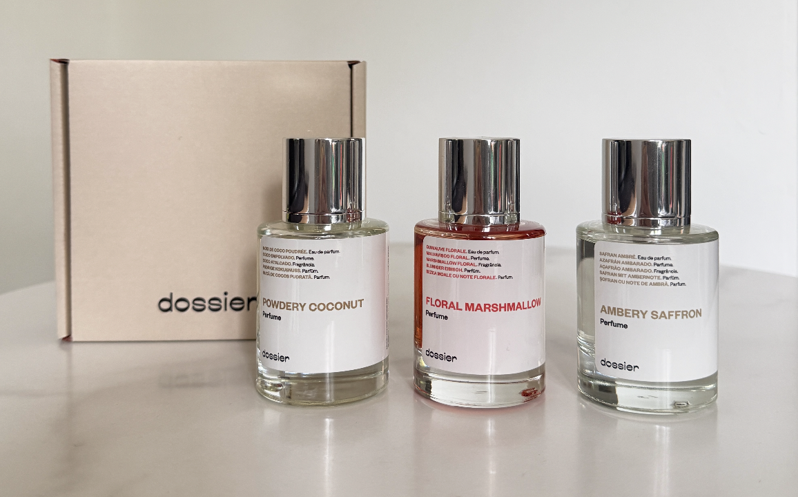 Photo of 3 Dossier Perfumes, "Powdery Coconut", "Floral Marshmallow" and "Ambery Saffron" pictured on a white marble table. Square Dossier box also pictured.