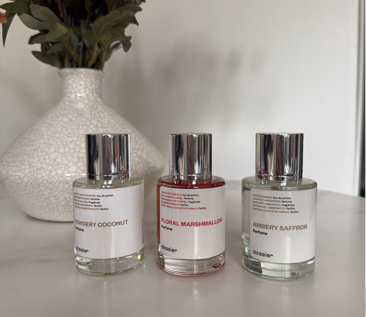 Photo of 3 Dossier Perfumes, "Powdery Coconut", "Floral Marshmallow" and "Ambery Saffron" pictured on a white marble table