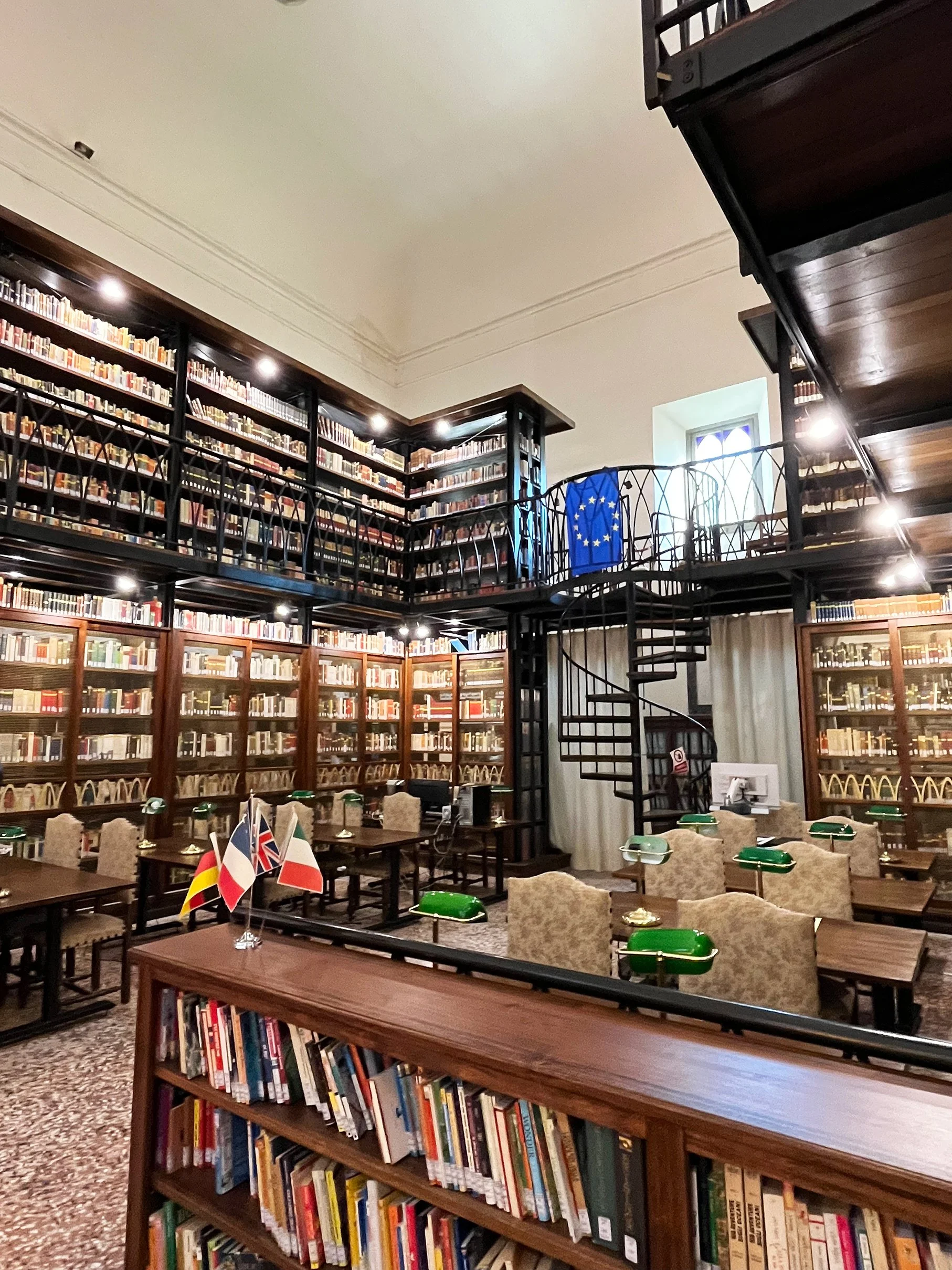 double storey dark bookshelves with a spiral staircase; dark wooden desks with green lamps