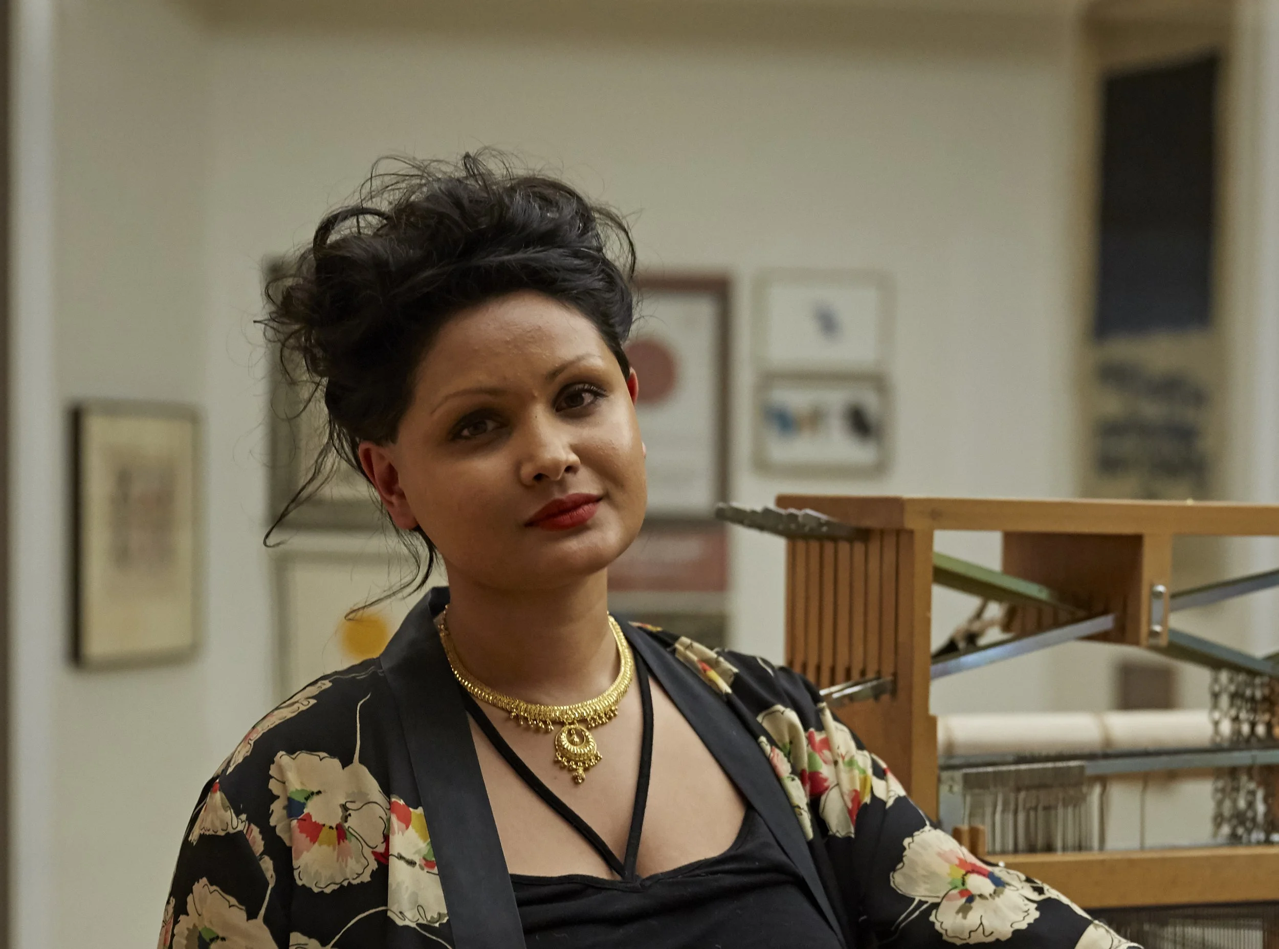 Portrait of artist Raisa Kabir standing beside a wooden weaving loom in a gallery setting. She faces the camera with a relaxed expression and wears a black top with a patterned floral jacket and a gold necklace. The background includes framed artwork