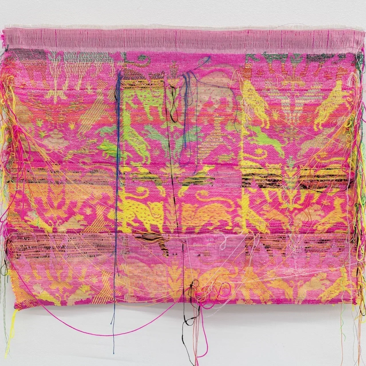 A rectangular woven textile in bright pink, yellow and green hangs on a white wall. Symmetrical animal and floral motifs run across the surface, interrupted by horizontal cuts and loose threads that expose the fabric’s layered structure.