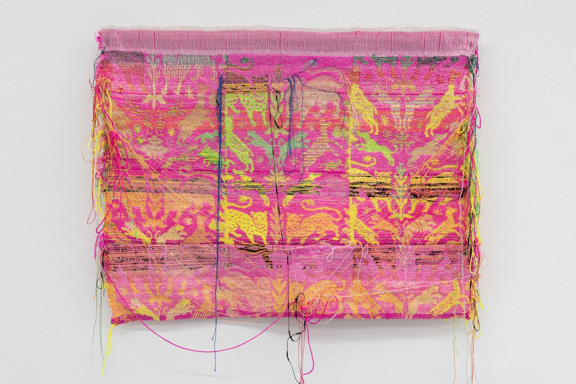 A rectangular woven textile in bright pink, yellow and green hangs on a white wall. Symmetrical animal and floral motifs run across the surface, interrupted by horizontal cuts and loose threads that expose the fabric’s layered structure.