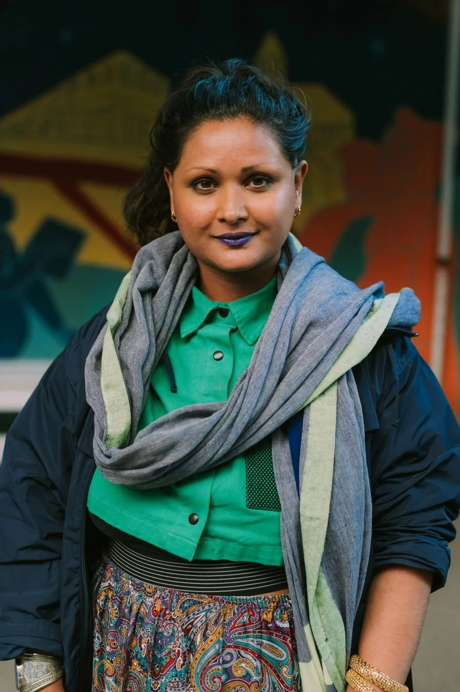 Portrait of artist Raisa Kabir standing outdoors wearing a green shirt, grey scarf and dark jacket.
