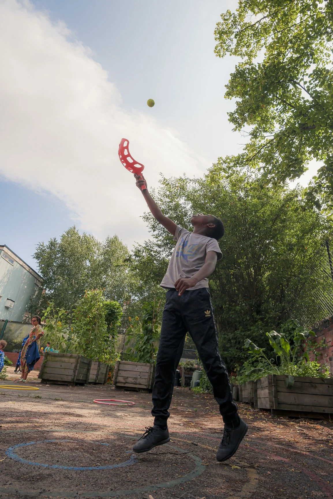 A young person jumps to catch a yellow ball in the air with a red plastic racket on a sunny day in a playground.