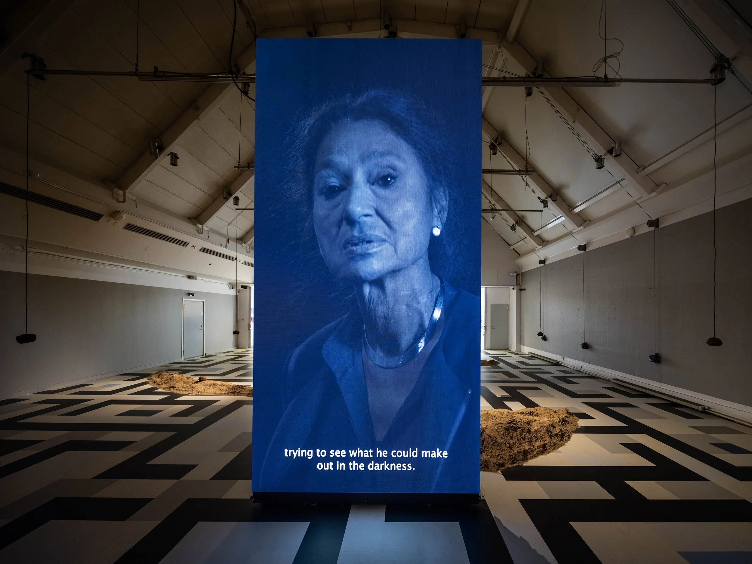 Blue-toned video portrait of a woman projected on a tall screen inside a gallery space with patterned flooring and small mounds of earth.