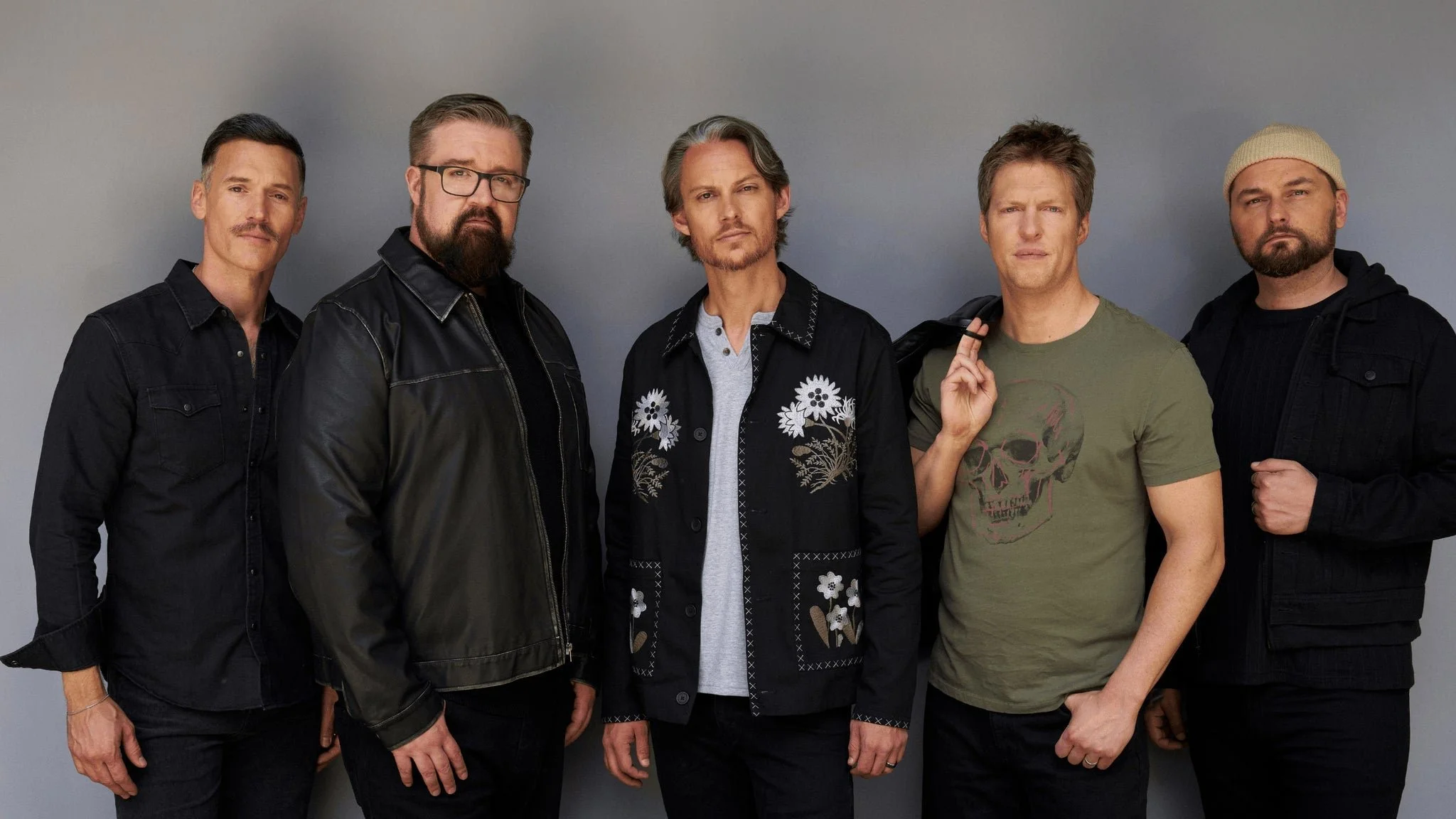 Group of five men standing against a gray background, dressed in casual and dark clothing, with serious expressions.