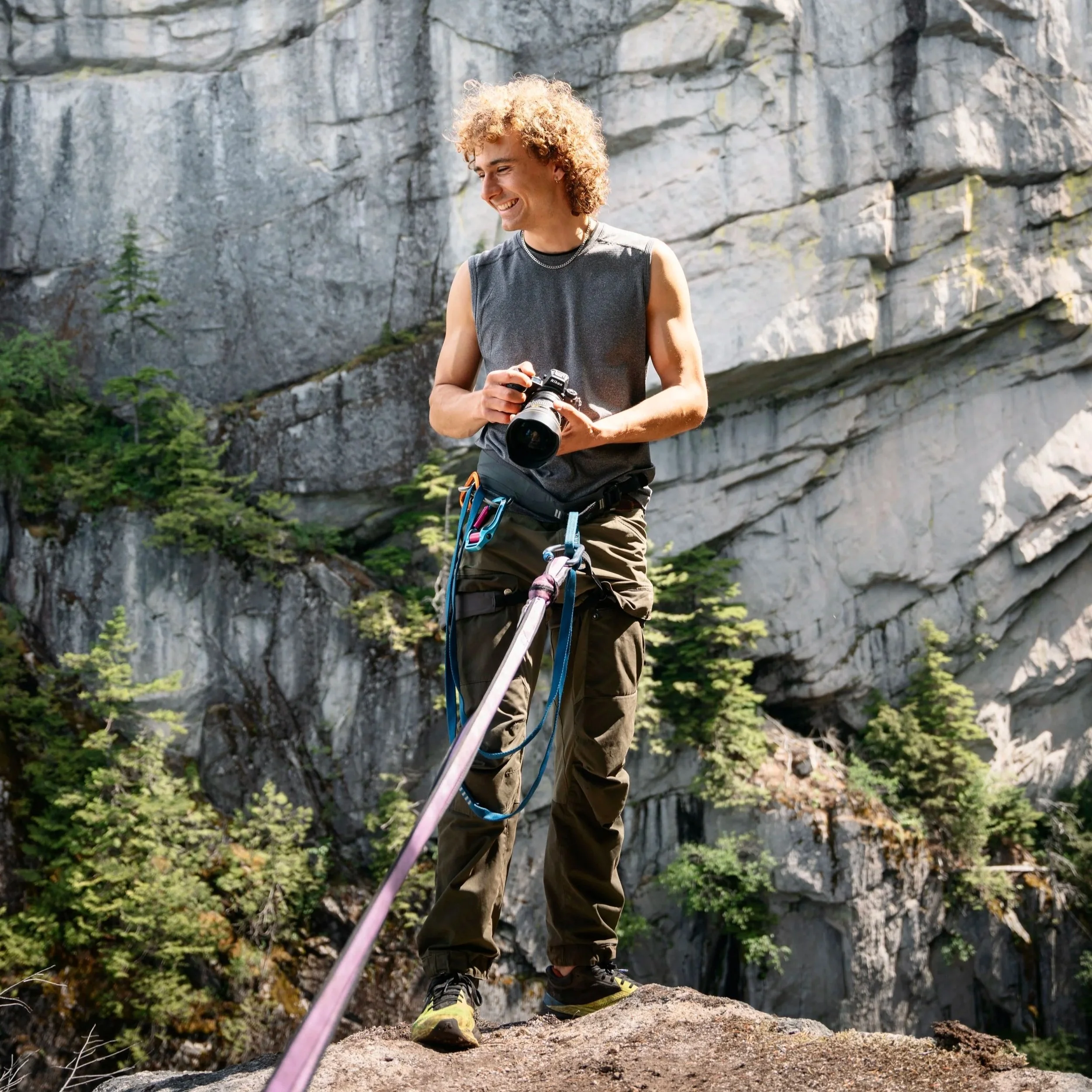 A young man with curly blonde hair wearing a sleeveless shirt and climbing gear stands on a rock ledge outdoors with a steep, rocky cliff in the background. He is smiling and holding a camera.