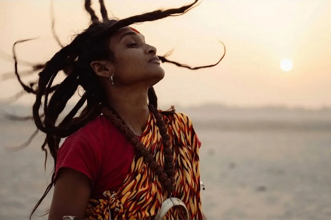 A woman with dreadlocks and traditional clothing standing by the water at sunset.