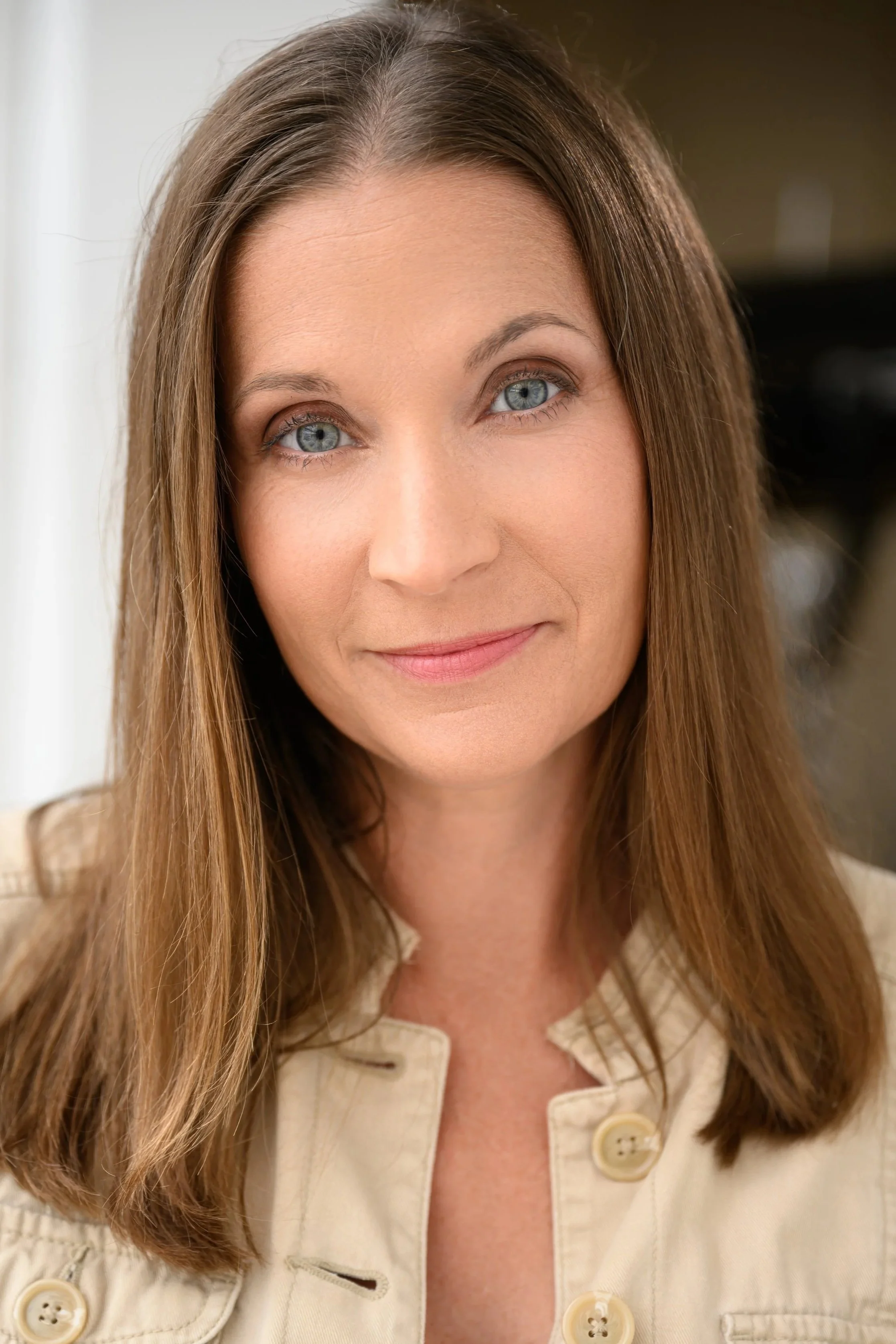 Close-up of a woman with long brown hair and blue eyes, wearing a cream-colored button-up shirt, smiling gently.