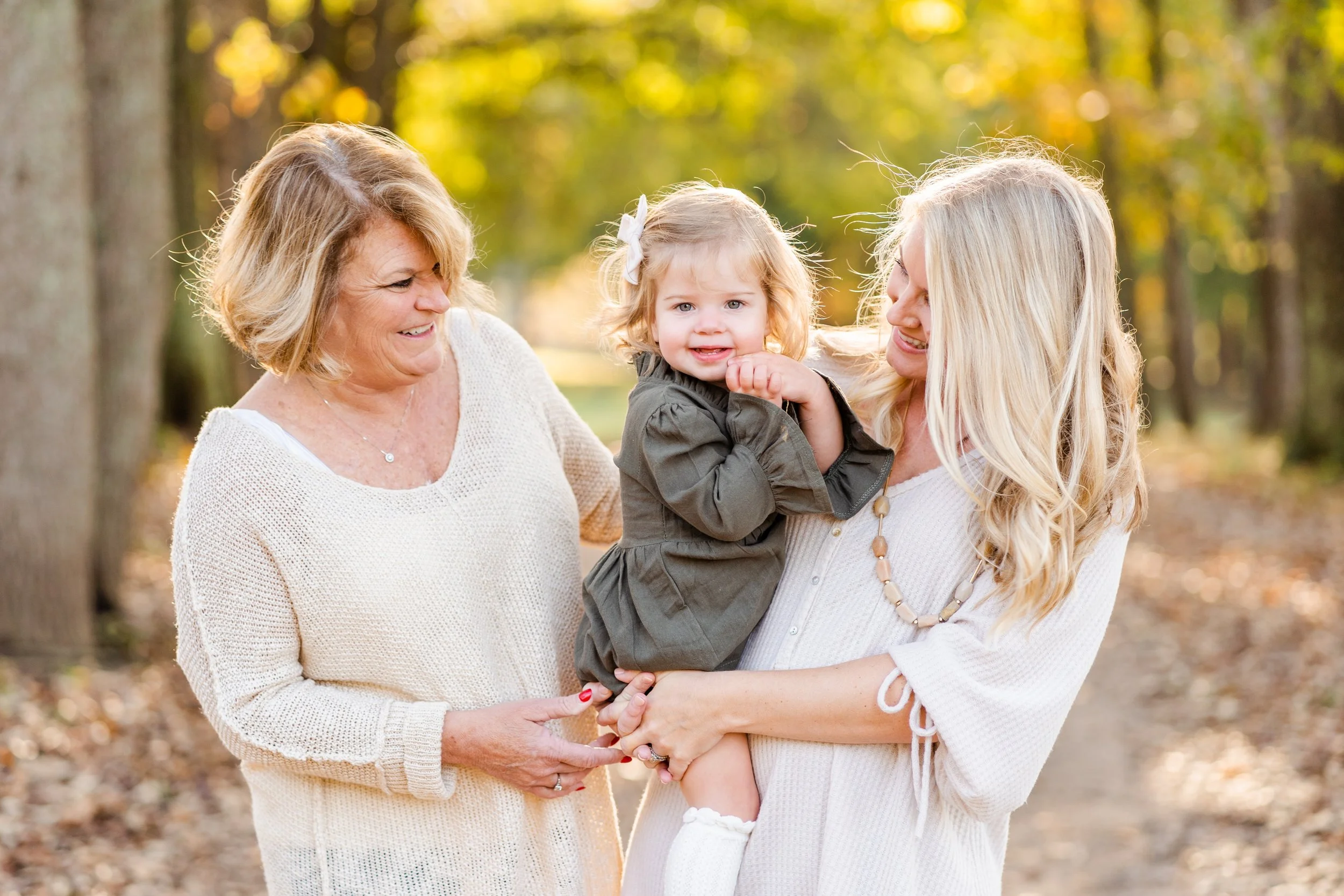 Warm fall family mini session with mother holding her smiling toddler and grandmother on opposite side smiling at grandaughter with fall trees and fall leaves in the background