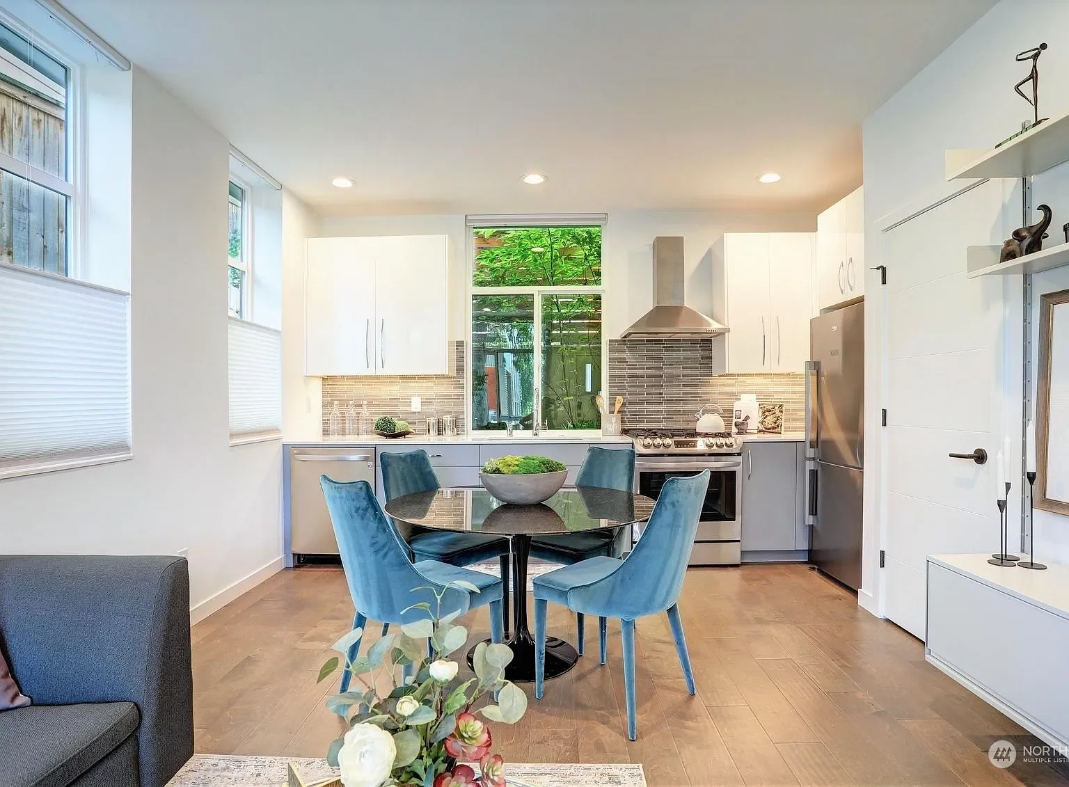 Modern kitchen with white cabinets, stainless steel refrigerator, gray stove and vent hood, blue dining chairs around a round glass table, wood flooring, green plants outside window.
