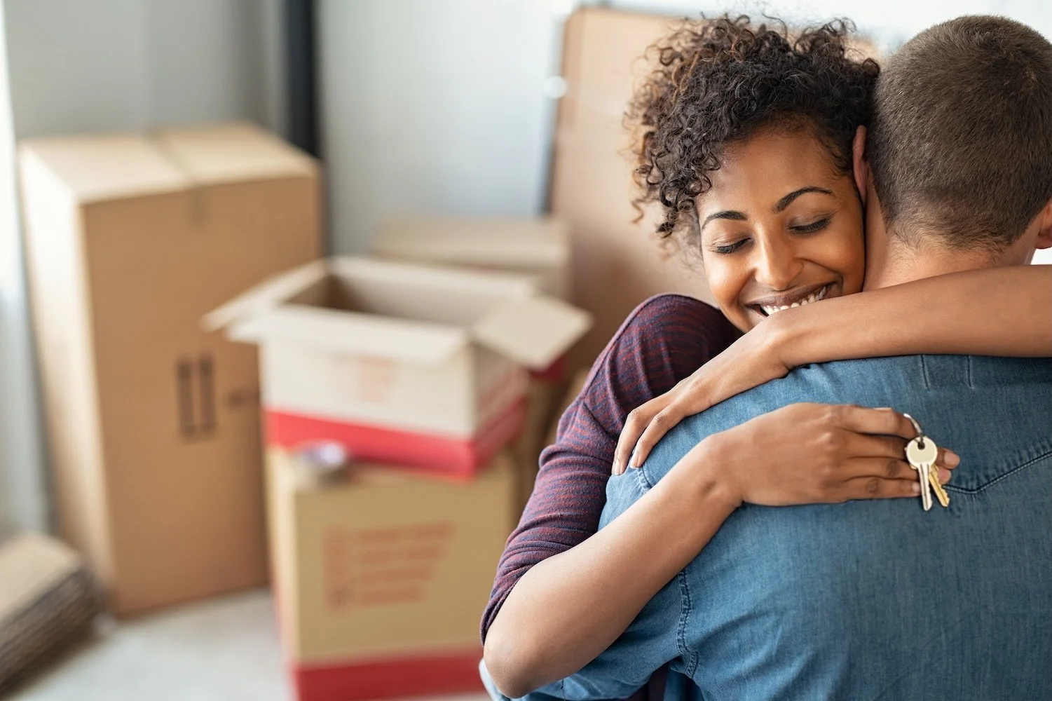 A woman smiling happily as she embraces a man, holding keys in her hand, in an indoor setting with moving boxes in the background.