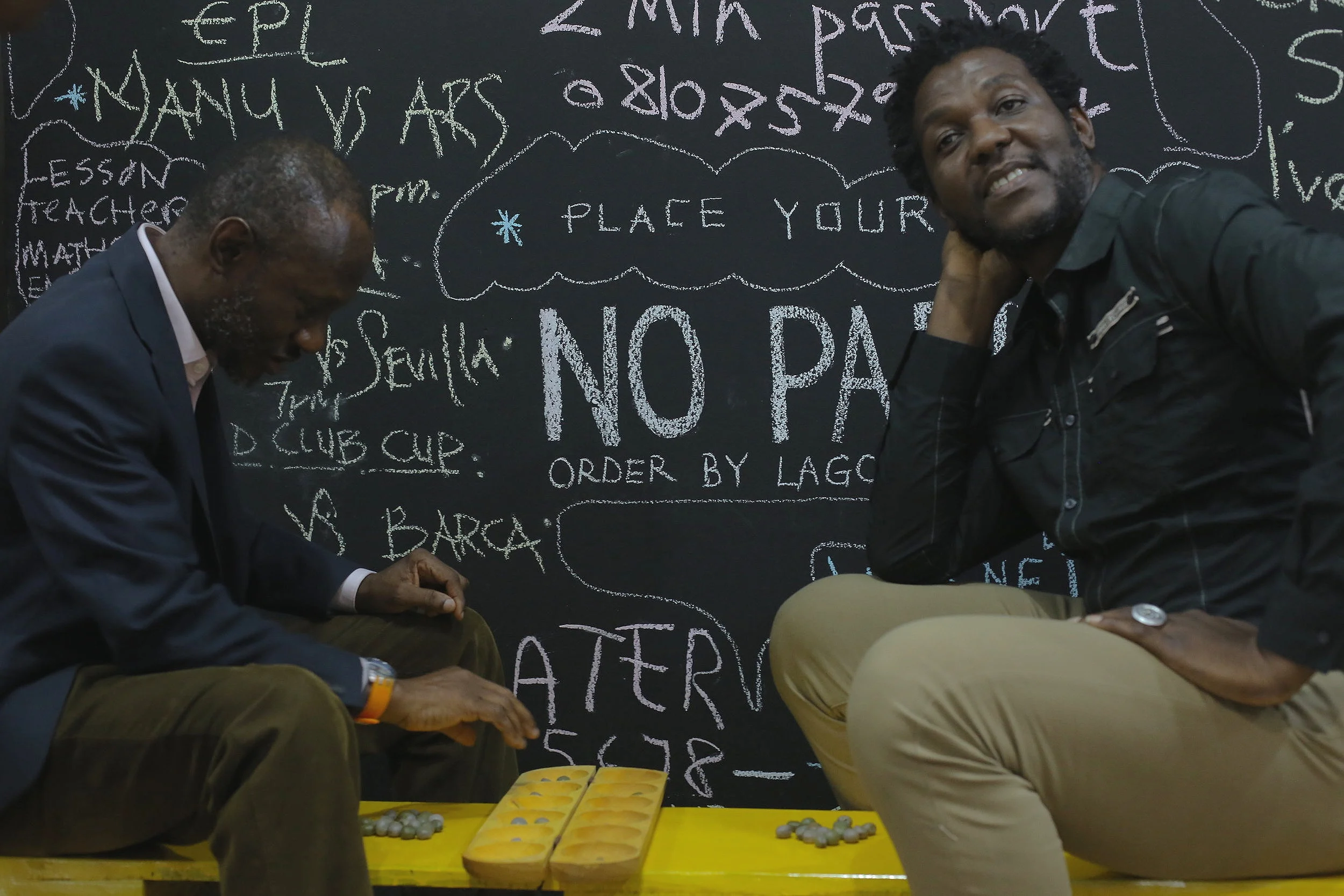 Conversationalists Aesthetics of the Wall Chuka Ihonor ( Architect and Urbanist) & Jude Idada ( Playwright and Film Maker) play a game of Ayo during the aesthetic of the wall experiment “In theatre, the wall is a space that separates the created reality from the actual reality." - Jude Idada ”When you watch a movie, you don't see an actor look directly at the camera (at you). Otherwise they're said to have broken the wall, the fourth wall. However, it could be intentional as in Kevin Spacey (as Frank Underwood) in House of Cards, saying for example, "How quickly poor grades are forgotten in the shadow of power and wealth'' into the camera (to pull you as audience into the created reality). The power of breaking the fourth wall is described as “the alienation effect”. It is a technique used to break down the comfortable distance between us, the audience watching unnoticed, and an actor on stage directing their actions towards us, distancing us from the artificiality of the performance. Occasional soliloquies were used by Shakespeare to address his audience in plays such as Hamlet, As You Like It and Henry V. English author Tom Brown highlights French director Jean-Luc Godard’s use of direct address in films such as Breathless, Week End and Pierrot le Fou”