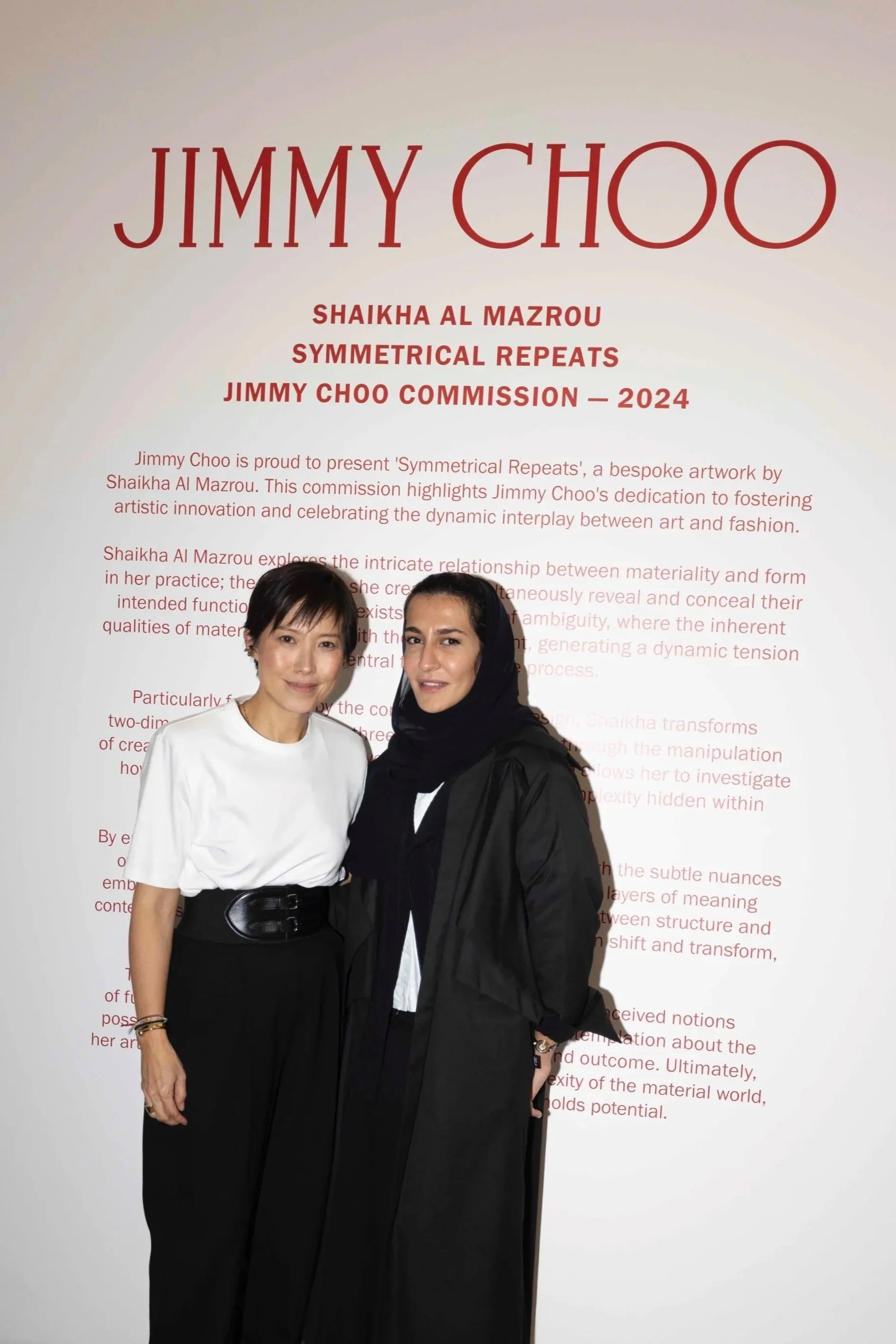 Two women standing in front of an art exhibition display titled 'Jimmy Choo' by Shaikha Al Mazrou, with text about subject matter.
