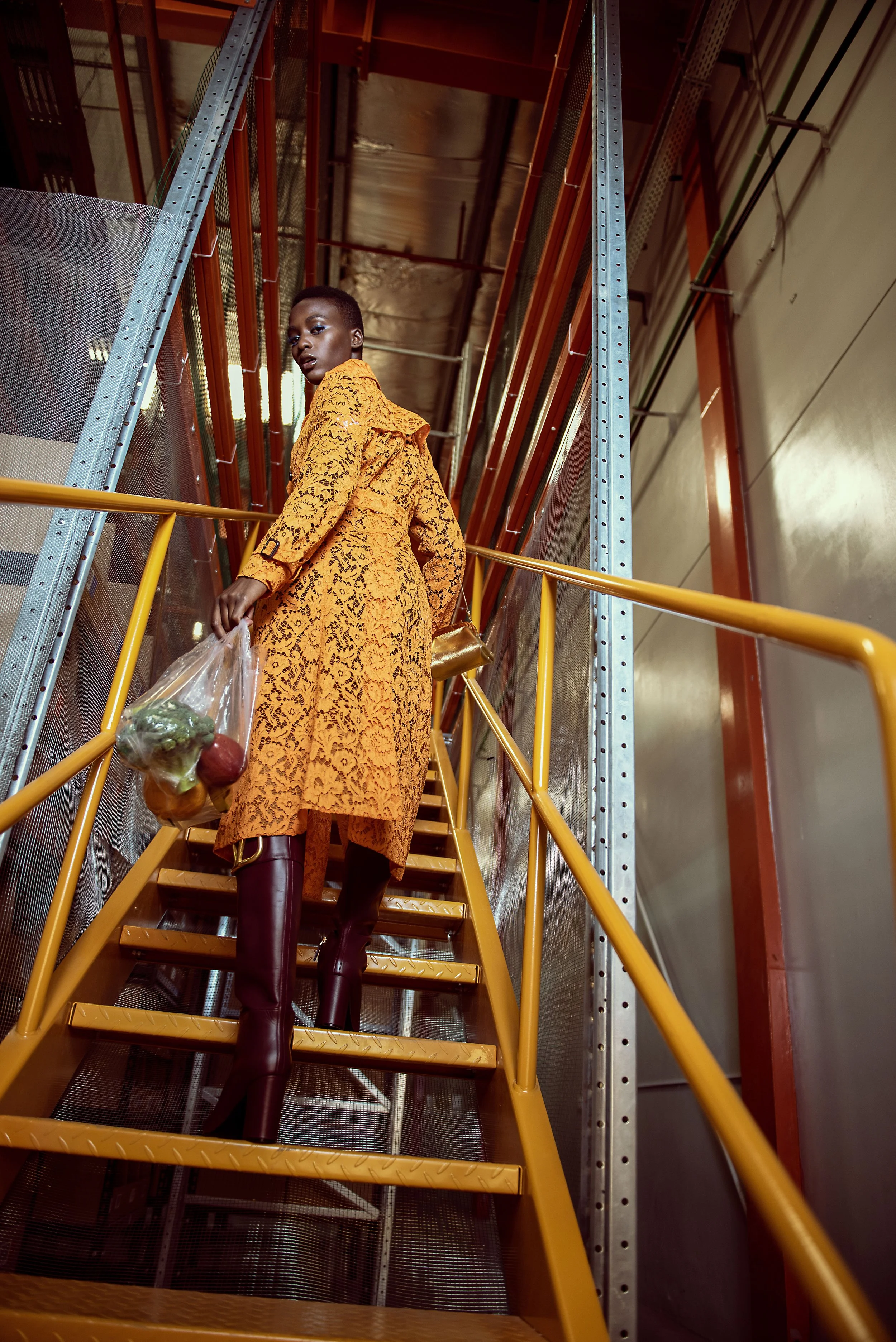 A woman in a yellow lace dress and matching boots holds a plastic bag of groceries while standing on a yellow staircase inside an industrial building.