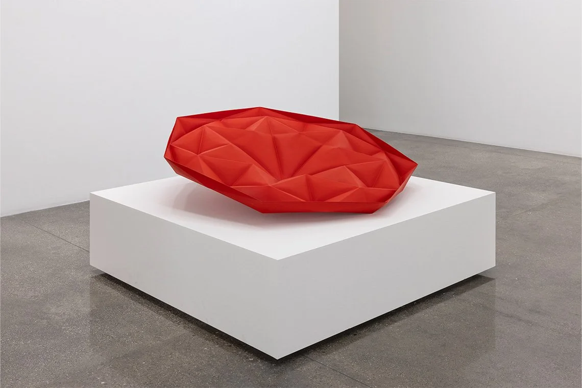 Red geometric sculpture on a white display pedestal in a modern art gallery.