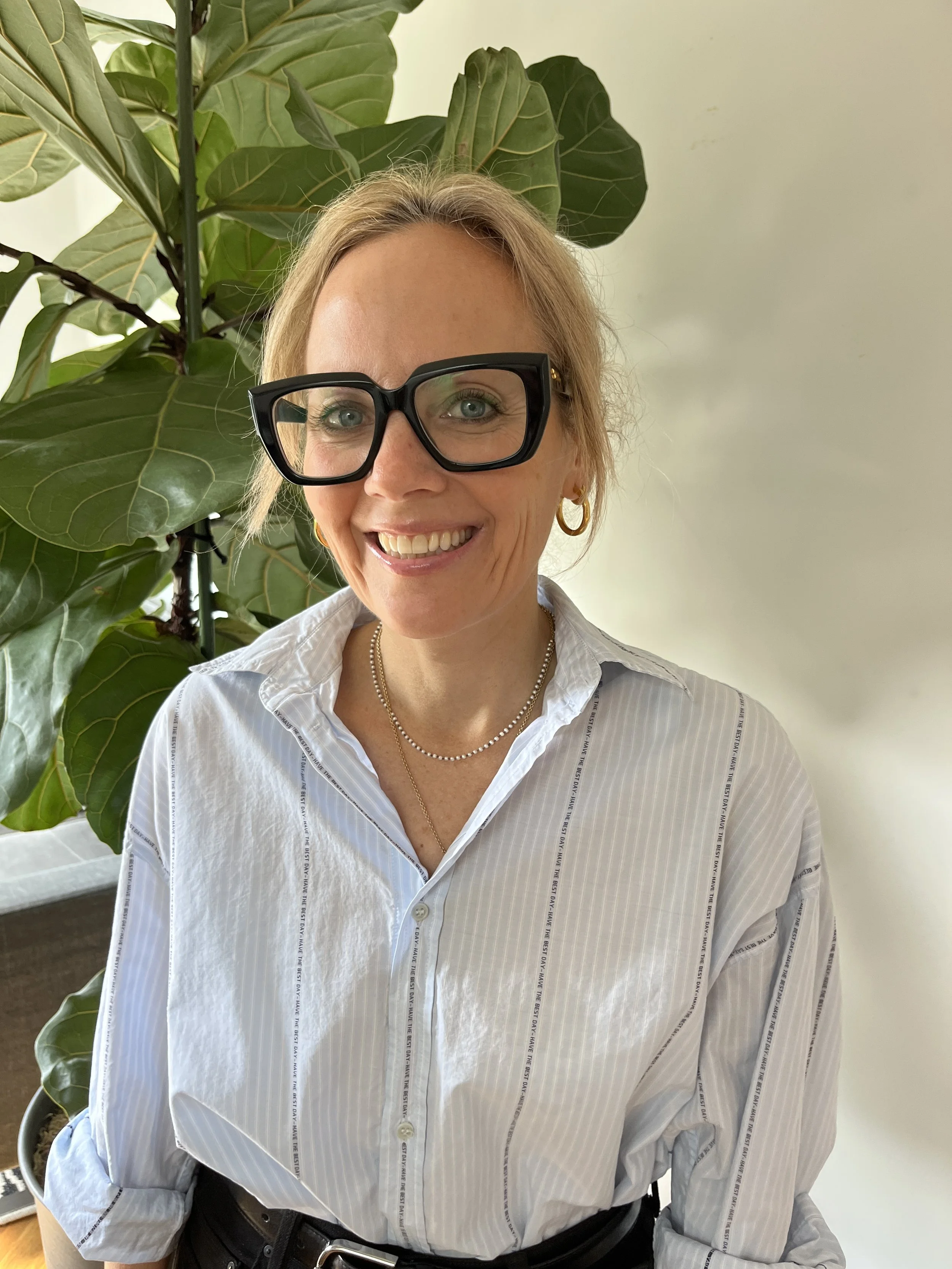 A woman with blonde hair wearing black glasses, gold hoop earrings, a pearl necklace, and a white striped shirt smiling in front of a large green leafy plant.
