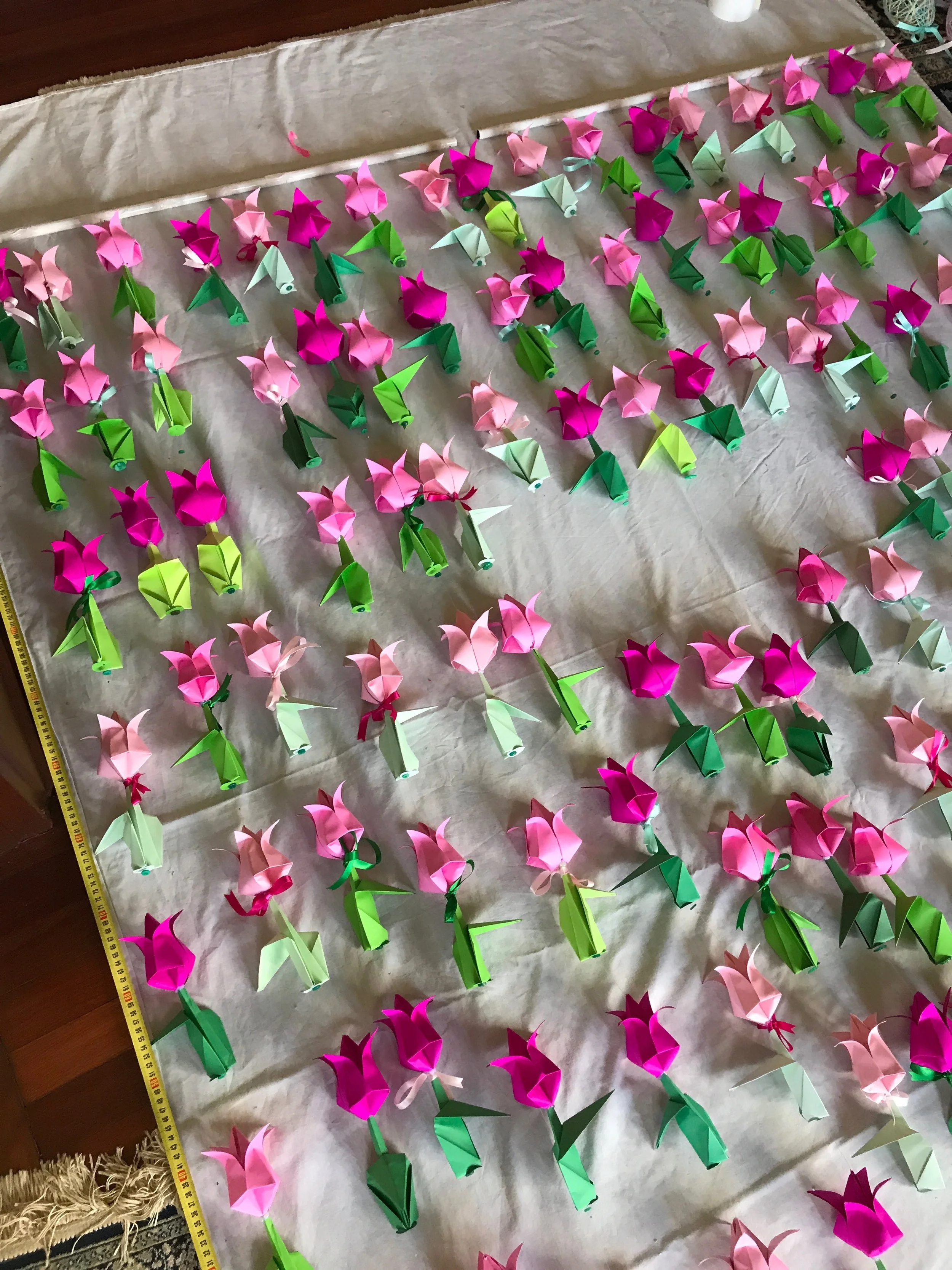 I measured out the window drop and arranged the tulips on a white sheet so I could get a good sense of the shapes in space. I then affixed small cardboard discs to the base of each tulip, in the same colour as the stems and threaded small green buttons below each tulip too, to allow them to sit flat and be strung in a line. I then secured each tulip using a tiny jewellery clamp.