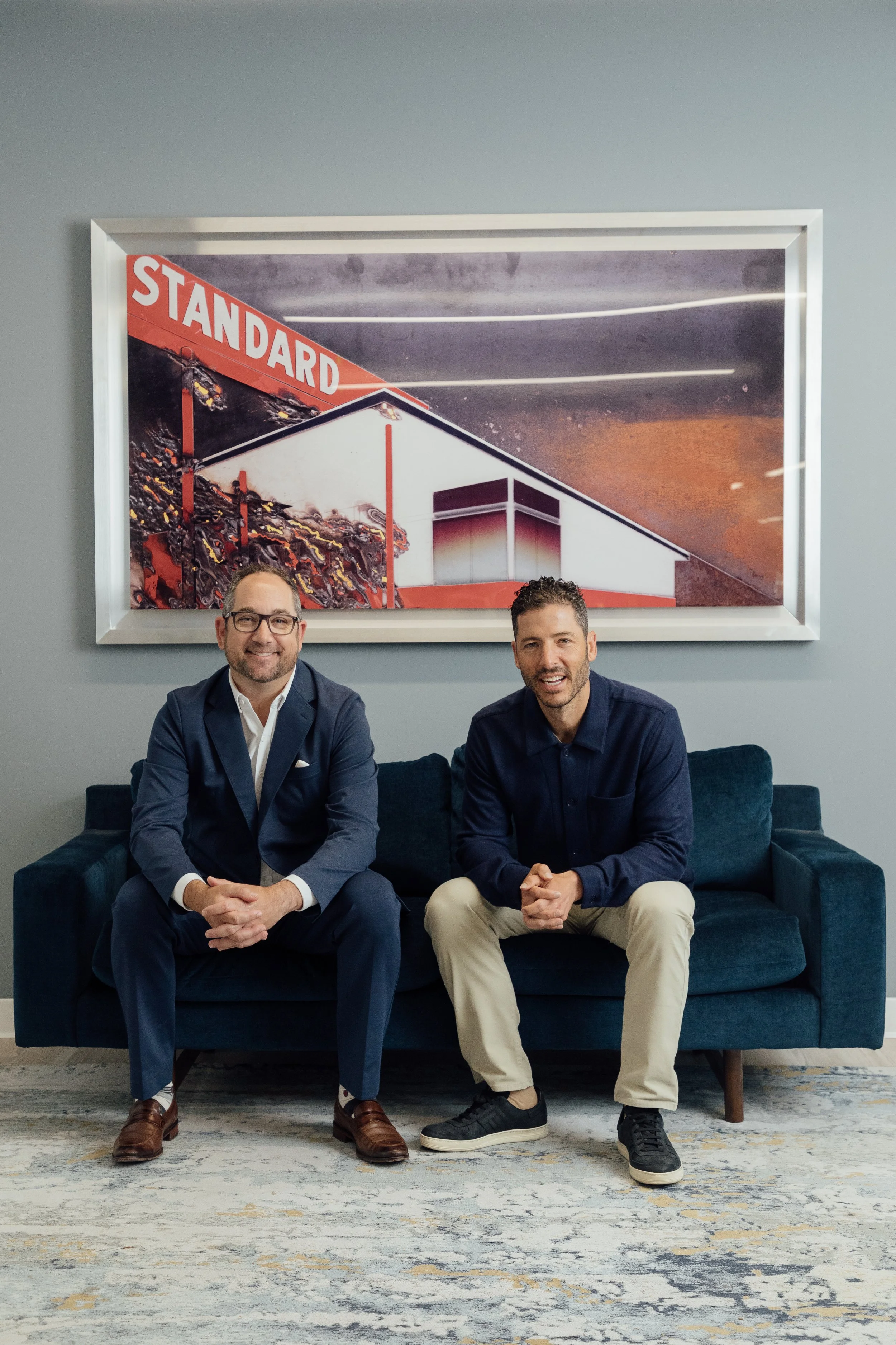 Two men sitting on a navy blue sofa in a modern office or lounge area. Behind them is a framed picture with the word 'STANDARD' and a building or structure in the background. The man on the left is wearing a blue suit, white shirt, and brown shoes. T