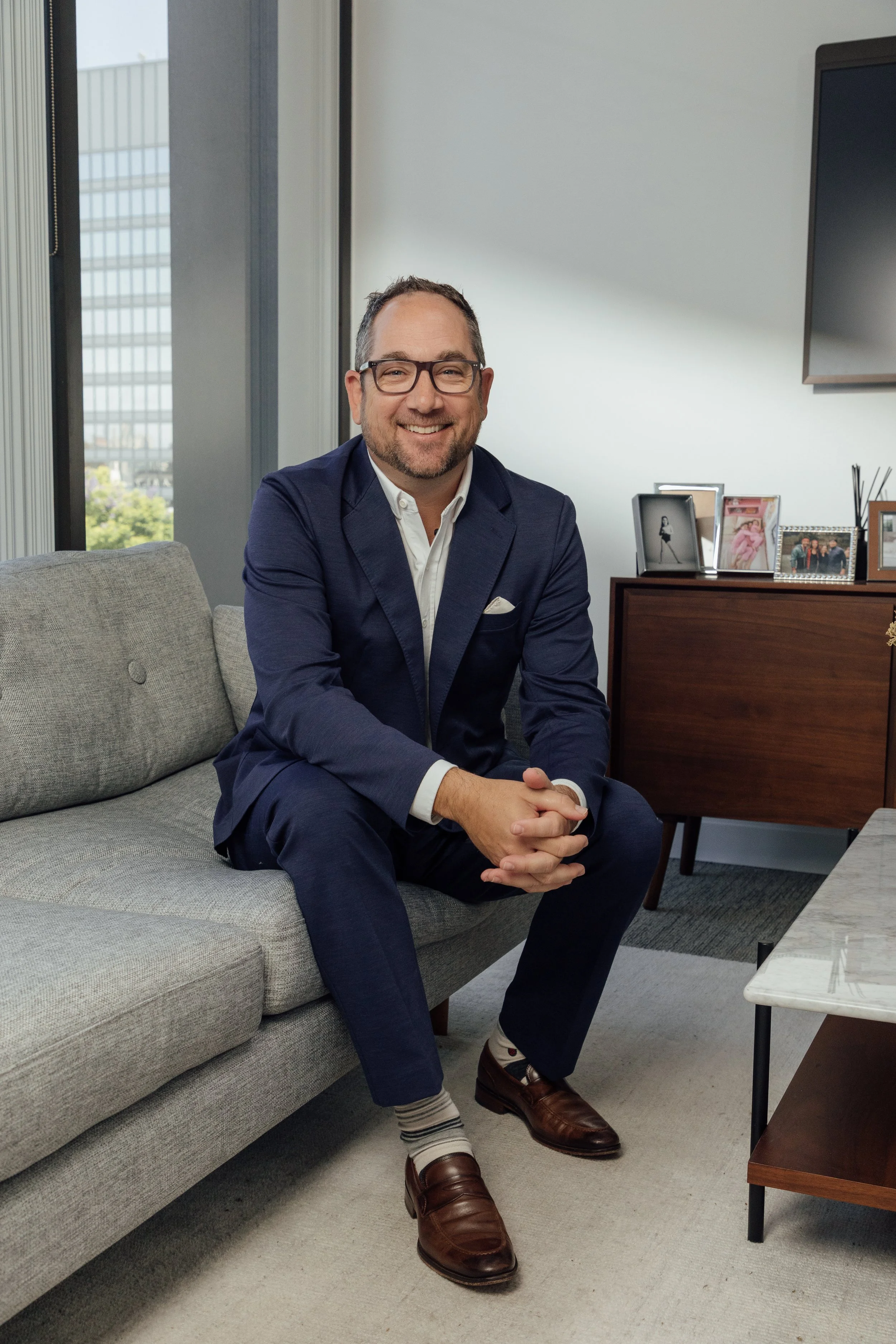 A man in a blue suit with glasses sitting on a gray sofa, smiling and clasping his hands, in an office with framed photos and a window with cityscape view.