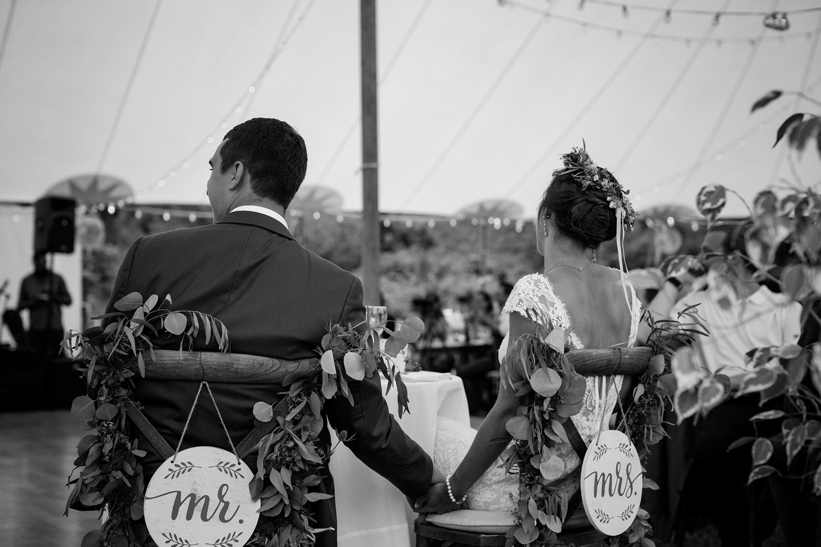 Bride and groom holding hands at their reception sweetheart table with cross back wooden chairs, greenery and mr. and mrs. signs hanging on the back of their chairs - Pearl Weddings & Events