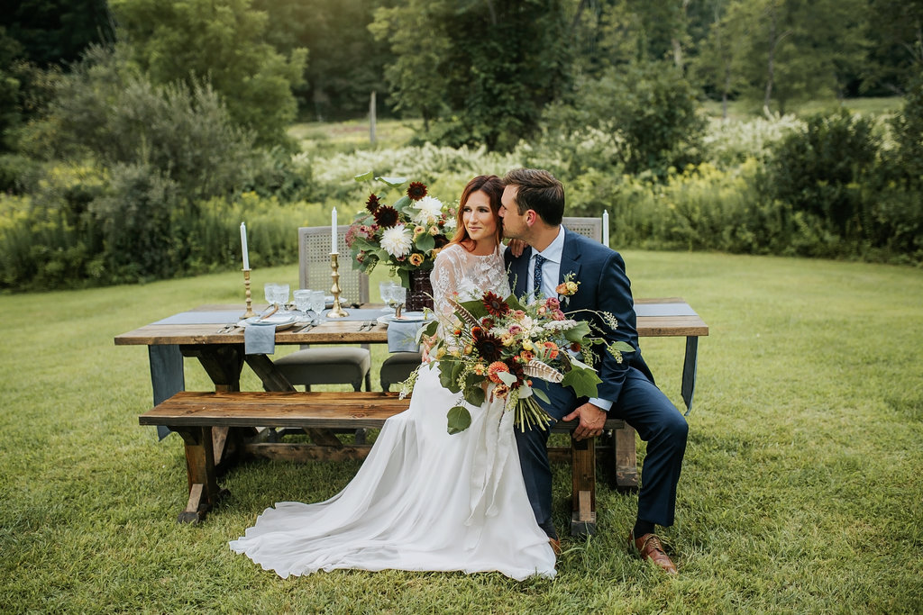 Bride and Groom at their farm table, bench and wicker chairs,  with blues and oranges hues at Great Hollow Nature Preserve -