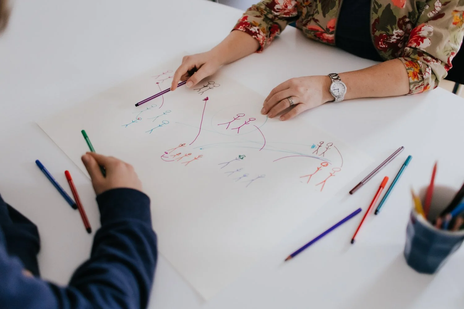 Twee mensen werken samen aan een schematische tekening met kleurrijke pennen op een witte tafel, met een pot met extra pennen ernaast.