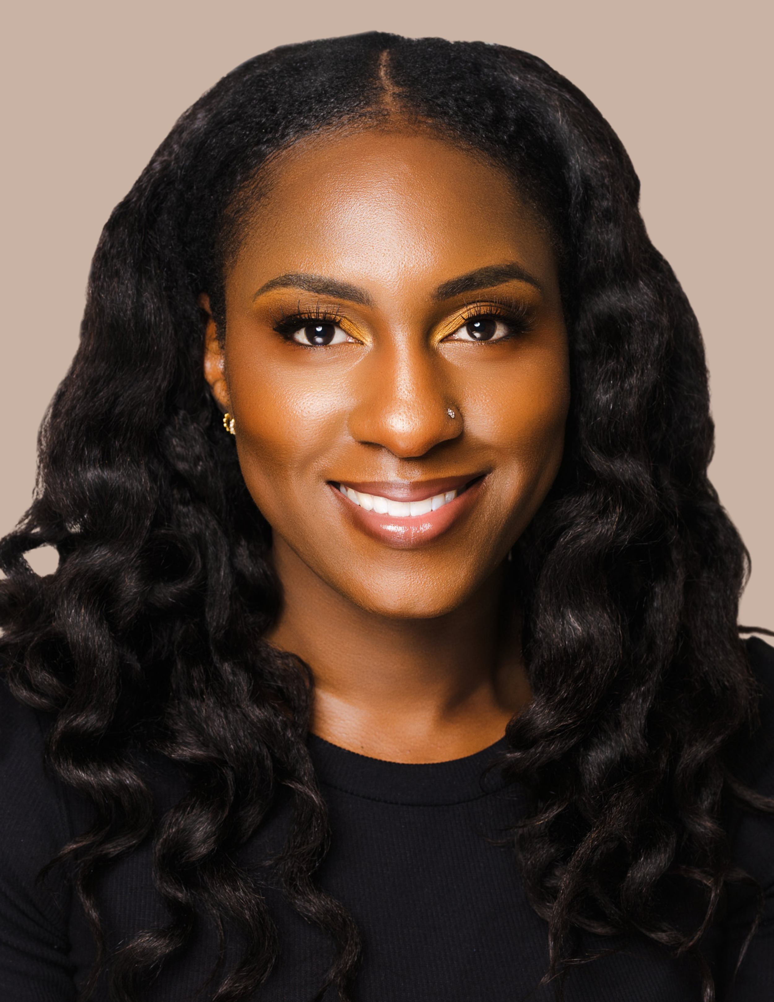 A woman with dark, curly hair smiling, wearing a black top and gold earrings, against a beige background.