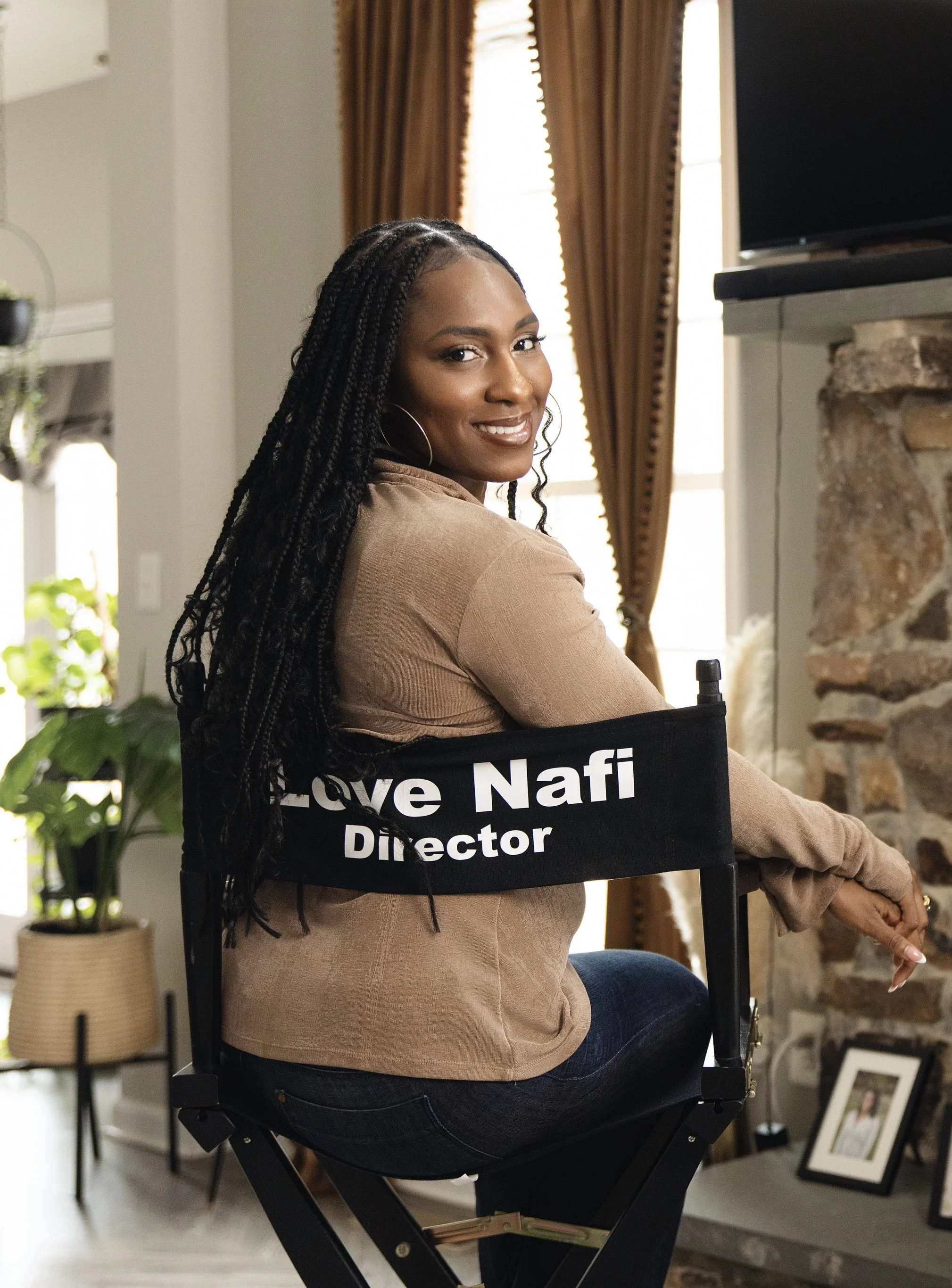A woman sitting on a director's chair with a sign that reads "Love Nafi Director". She is smiling and looking back at the camera, with a cozy indoor background featuring a stone fireplace, curtains, and framed photos.