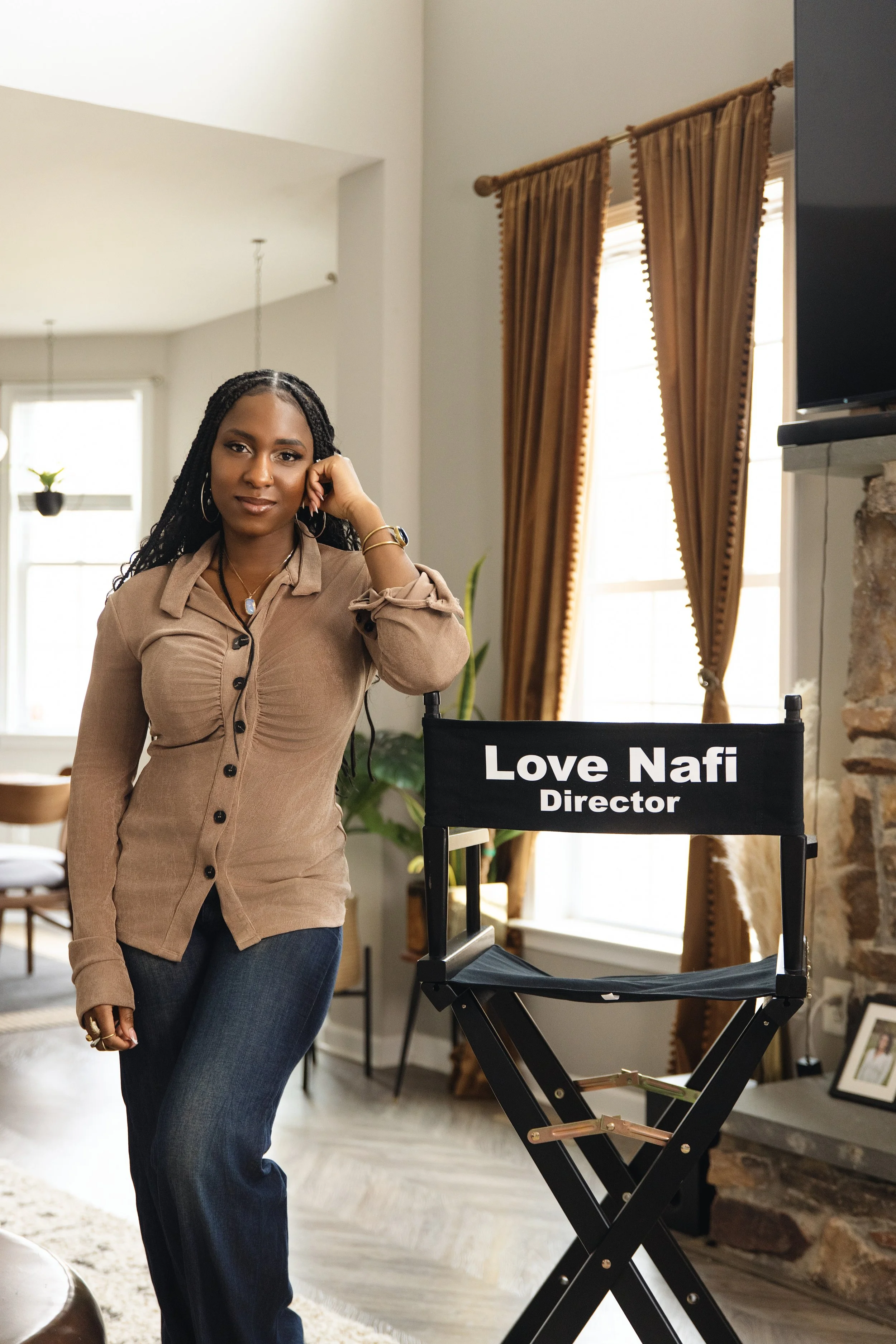 A woman standing indoors, wearing a beige button-up shirt and blue jeans, leaning on her left hand, with a director's chair labeled 'Love Nafi Director' nearby, and a window with brown curtains in the background.