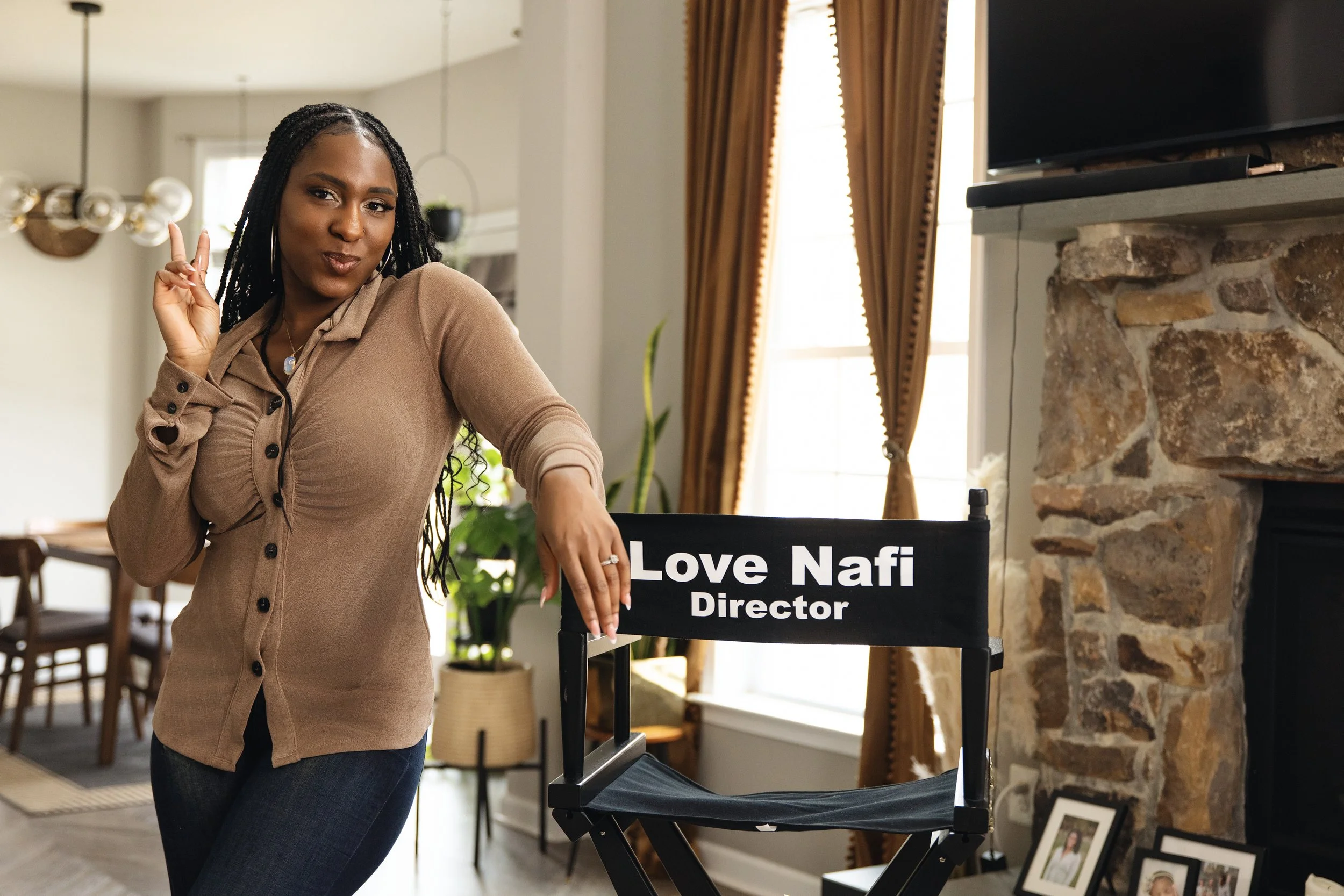 A woman with braided hair making a peace sign with one hand while standing next to a director's chair that reads "Love Nafi Director" in a cozy, well-lit room with brick fireplace and gold curtains.