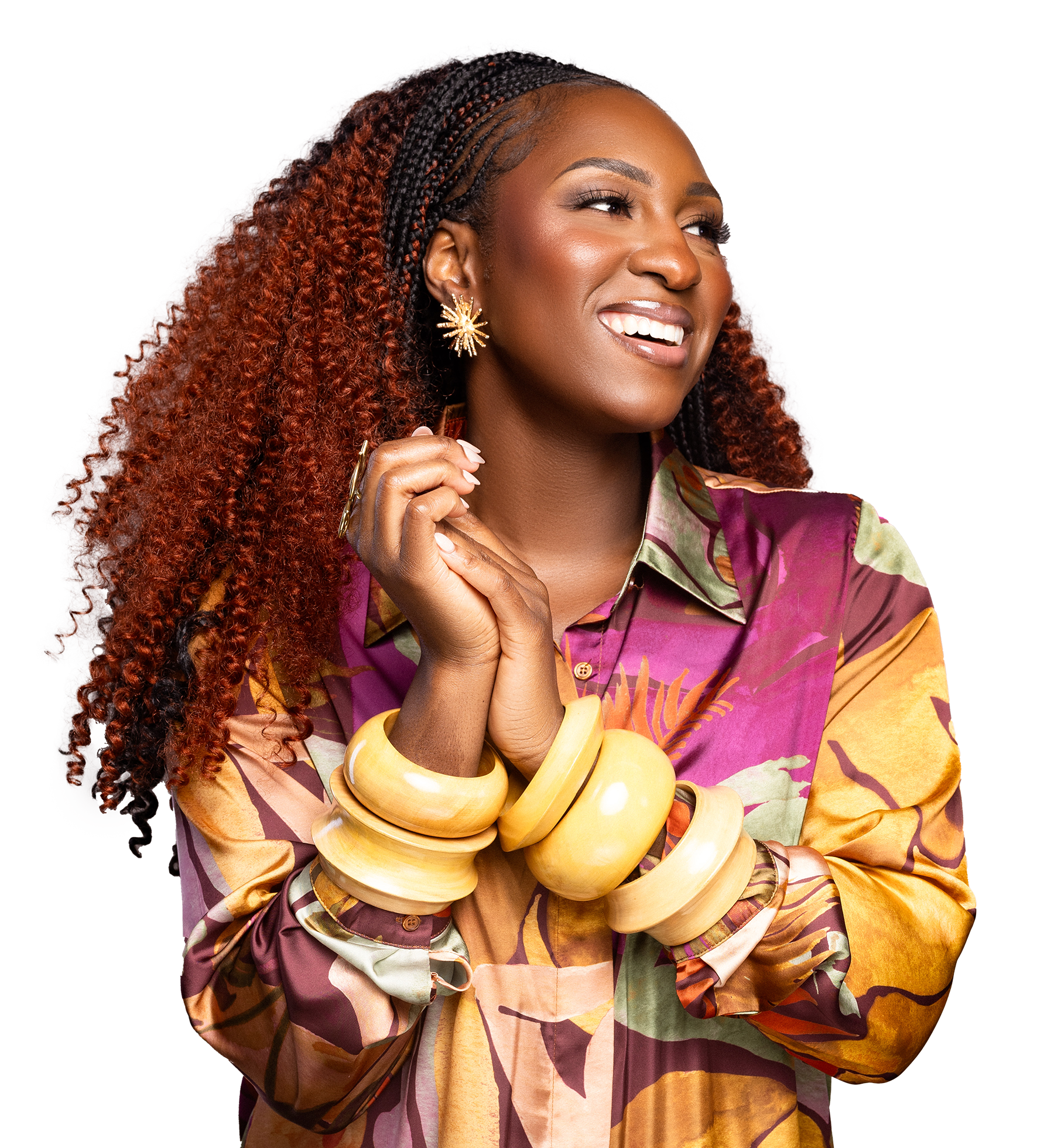 A woman with long curly hair styled in braids, wearing a colorful patterned shirt, large gold earrings, and multiple chunky yellow bracelets, smiling and looking to the side.