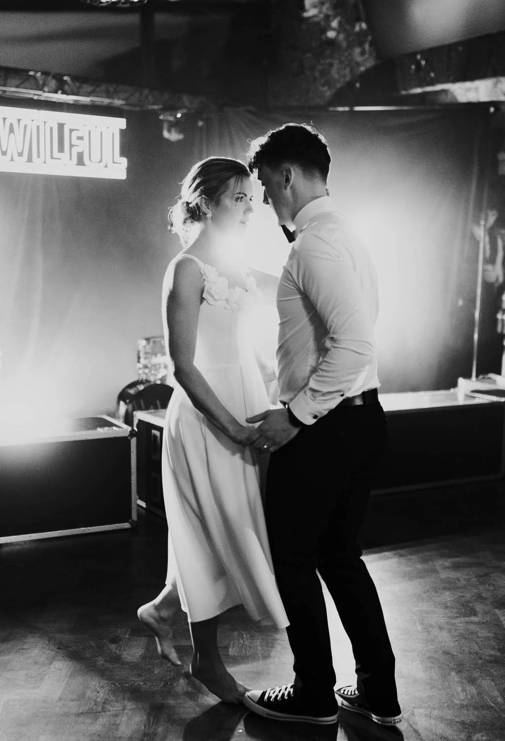 A black and white photograph of a couple dancing closely, the woman wearing a sleeveless dress and the man in a white shirt with a bow tie. They are holding hands and looking into each other's eyes, with a sign that reads 'WILFUL' visible in the back