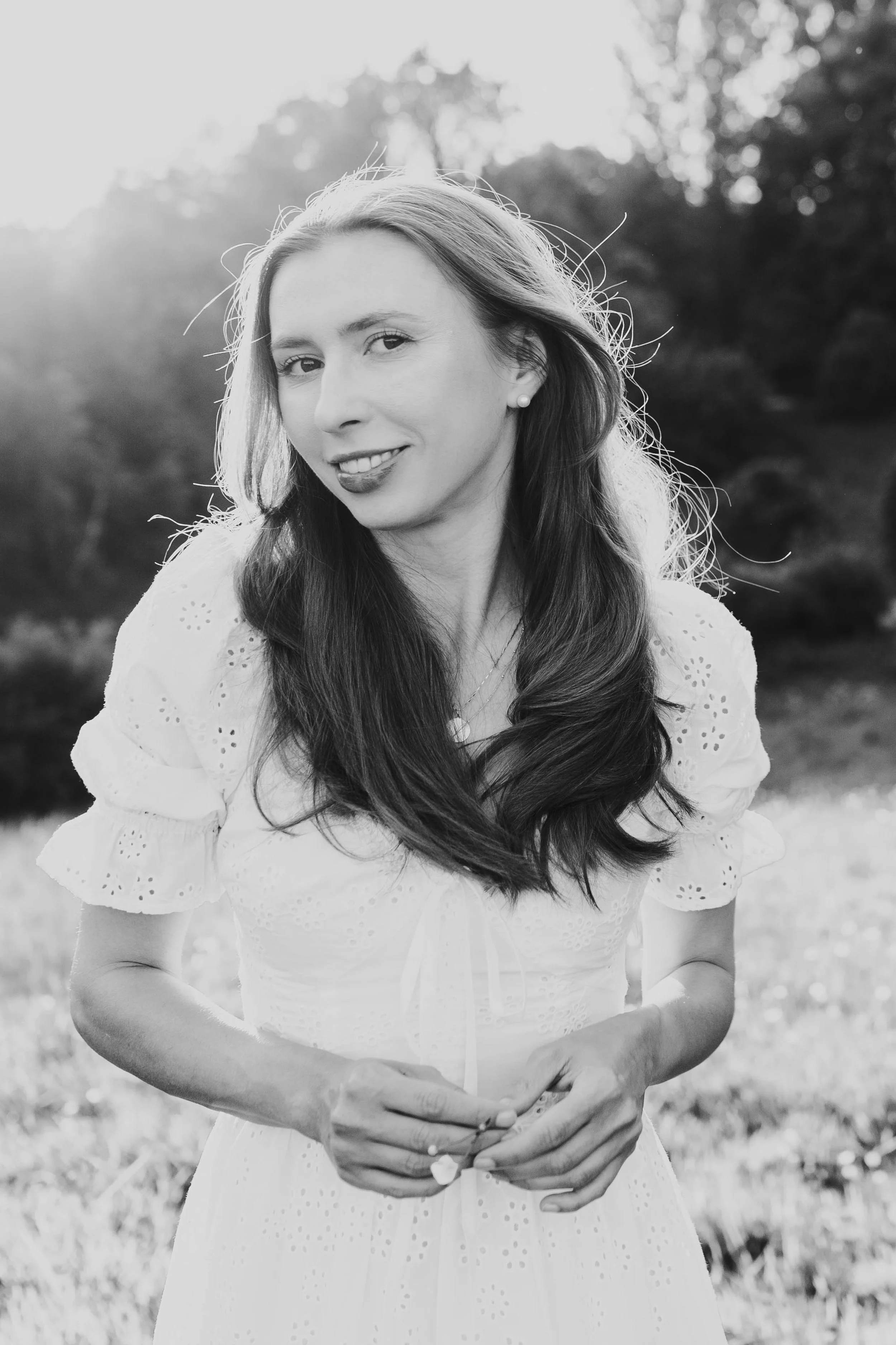 Black and white photo of a woman with long hair, smiling, wearing a dress with eyelet details, outdoors with trees in the background.