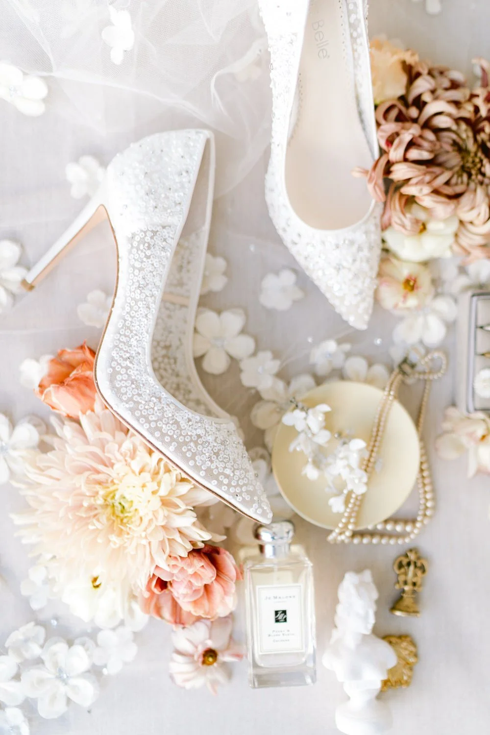 A pair of white high-heeled shoes with sequins, surrounded by soft pink and white flowers, a perfume bottle, a pearl necklace, and small decorative items on a light-colored surface.