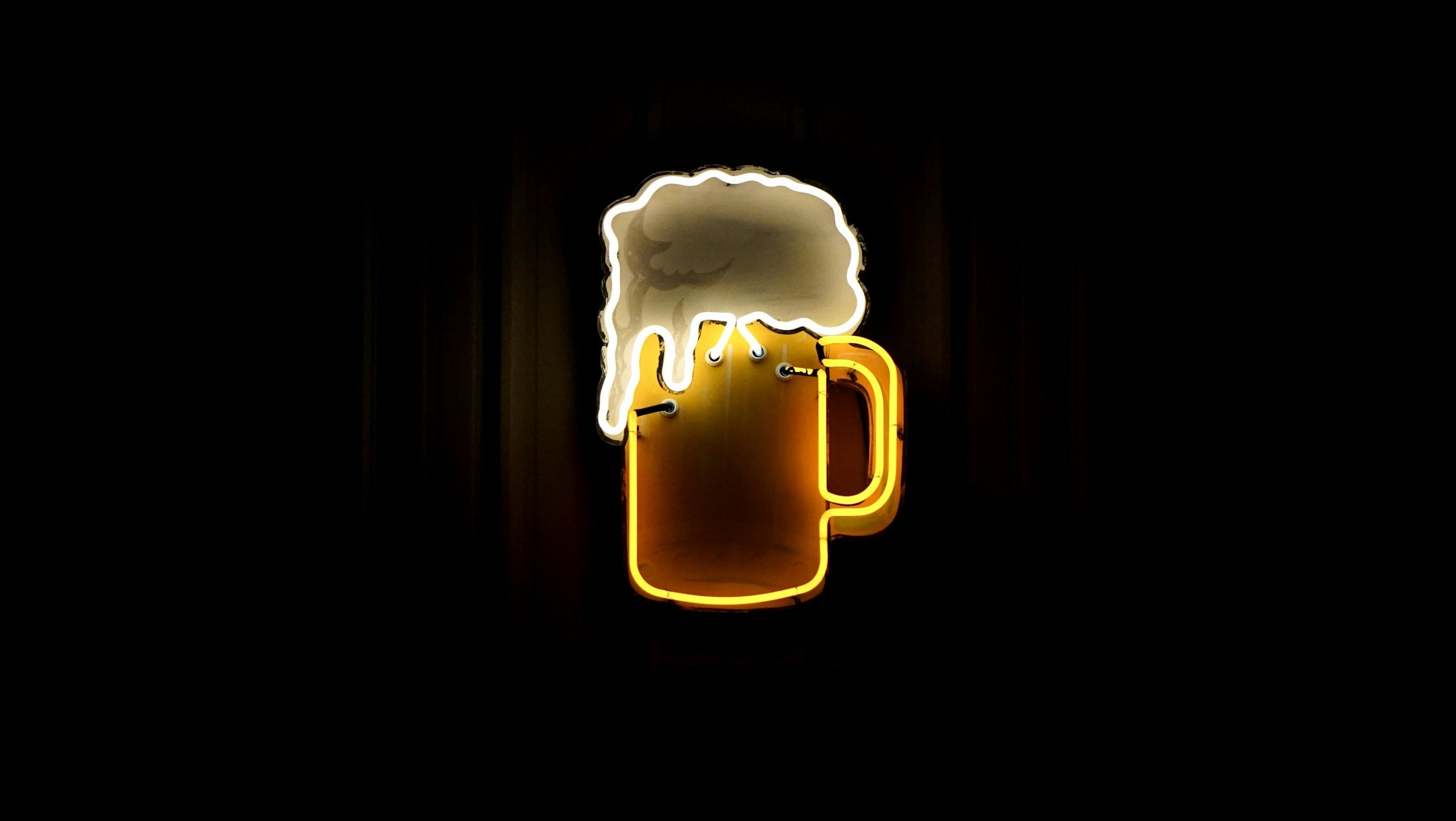 Neon sign of a beer mug with foam on top, glowing yellow and white colors against a dark background.