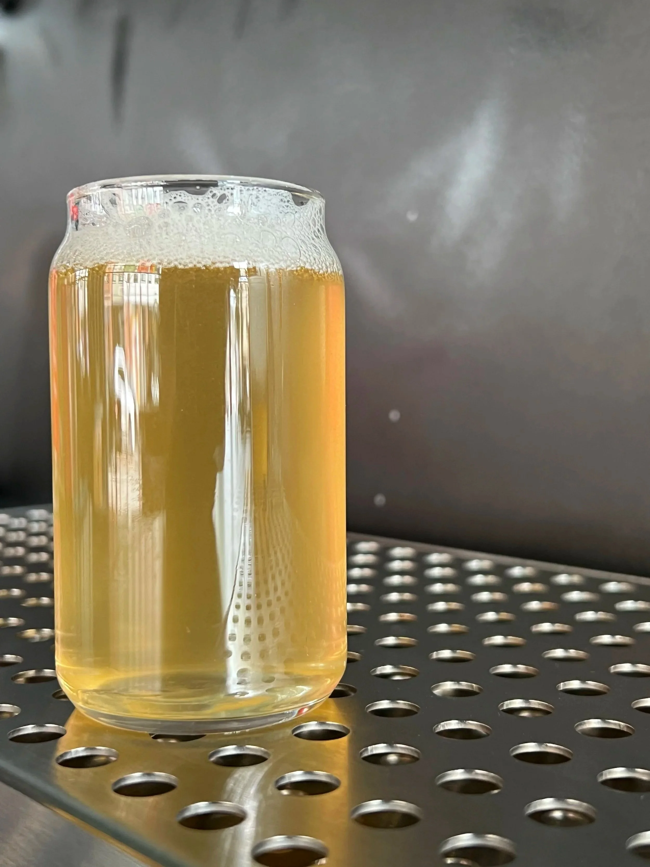 A glass of pale yellow beer with foam on top, placed on a black perforated metal surface in front of a dark background.