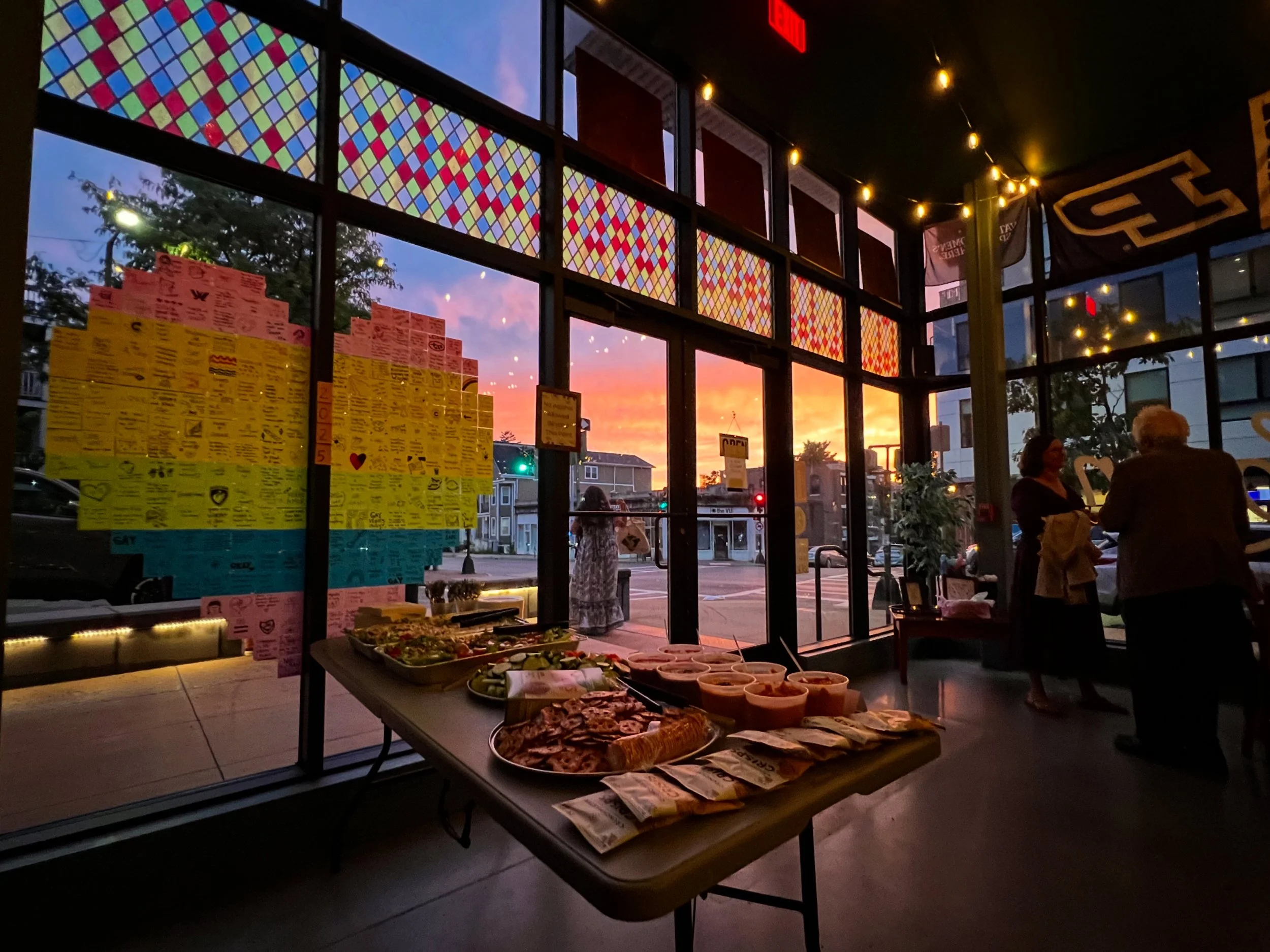 Indoor view of a restaurant during sunset with a table of food in the foreground and travelers outside in the street through large windows. Colorful stained glass adorns the top of the window. Private event food spread on table.