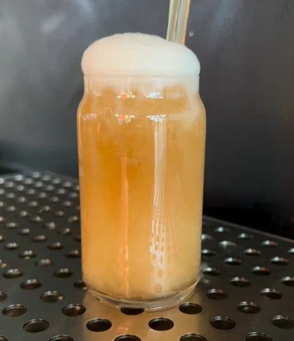 A glass of beer with foam on top and a straw, placed on a perforated metal surface.