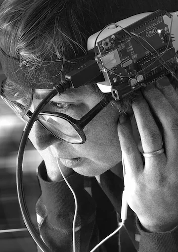 Image description: A person with short brown hair and glasses tilts their head downwards as they look off-screen. They are wearing an Arduino on a headband, their hands hold a pair of earbuds in their ears, and multiple wires extend downwards, out of the image. The image is grainy black & white, with the background blurred to remove extraneous information.
