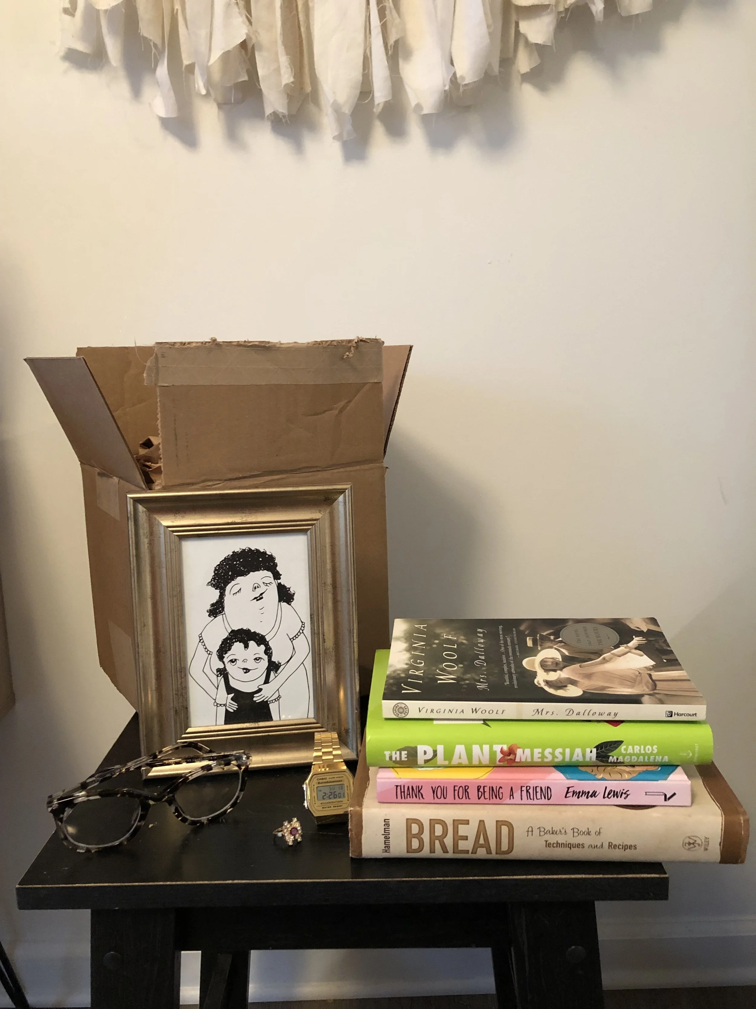 Image description: A stack of books, a pair of glasses, a ring, a watch, and a drawing sit on a chair in front of an open cardboard box, indicating sentimental objects included in one Home in a Box.