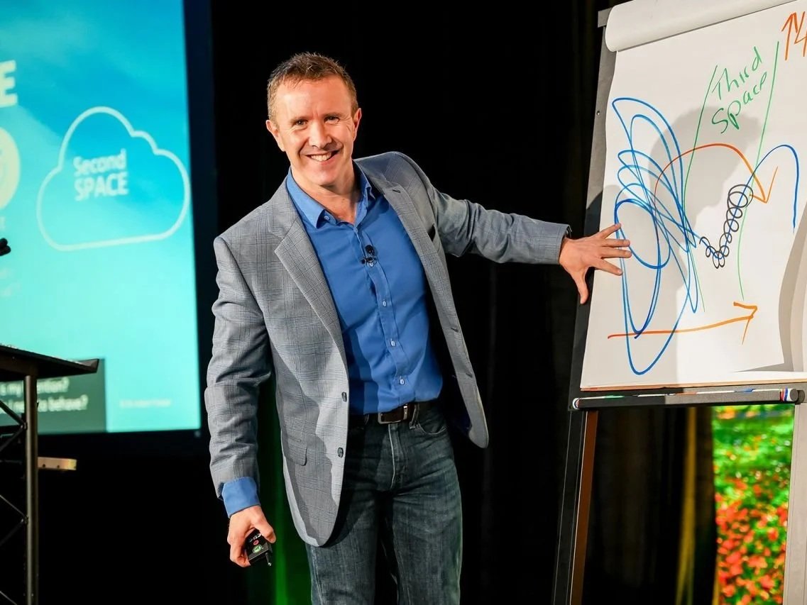 Dr Adam Fraser on stage presenting The Third Space keynote, smiling, with writing on a flipchart and a presentation on a large screen behind him