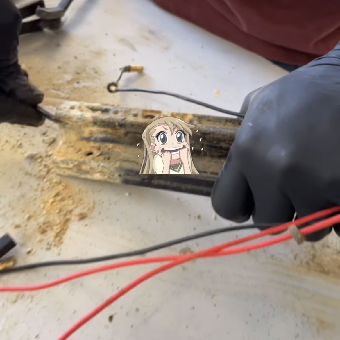 Una persona con guantes negros manipula cables sobre una superficie sucia, con un dibujo de un personaje animado superpuesto.