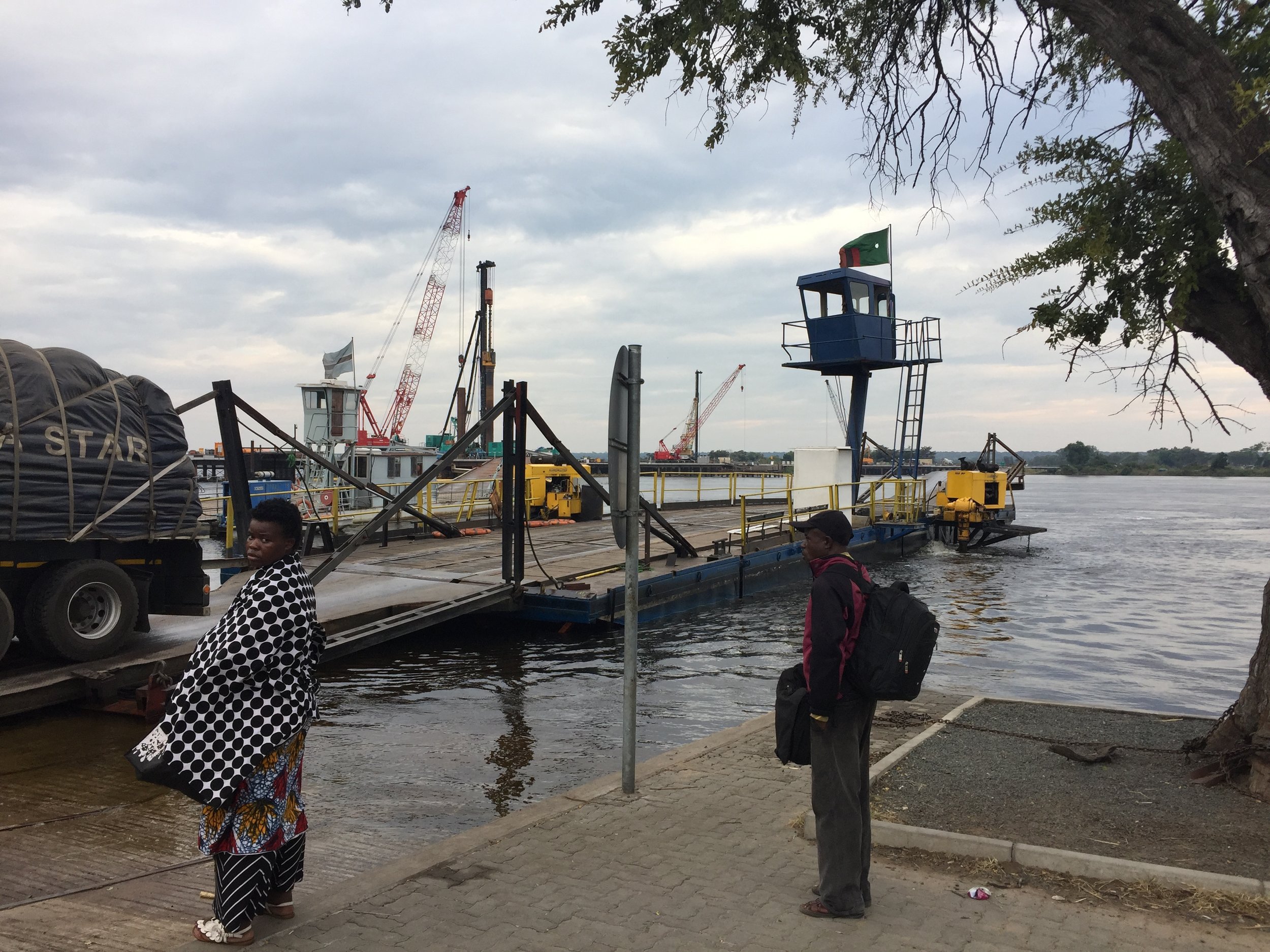 A woman wrapped in her floral dress and a blanket for the chilly morning waits alongside the Kazungula Ferry, while a big rig disembarks from the Zambia side.