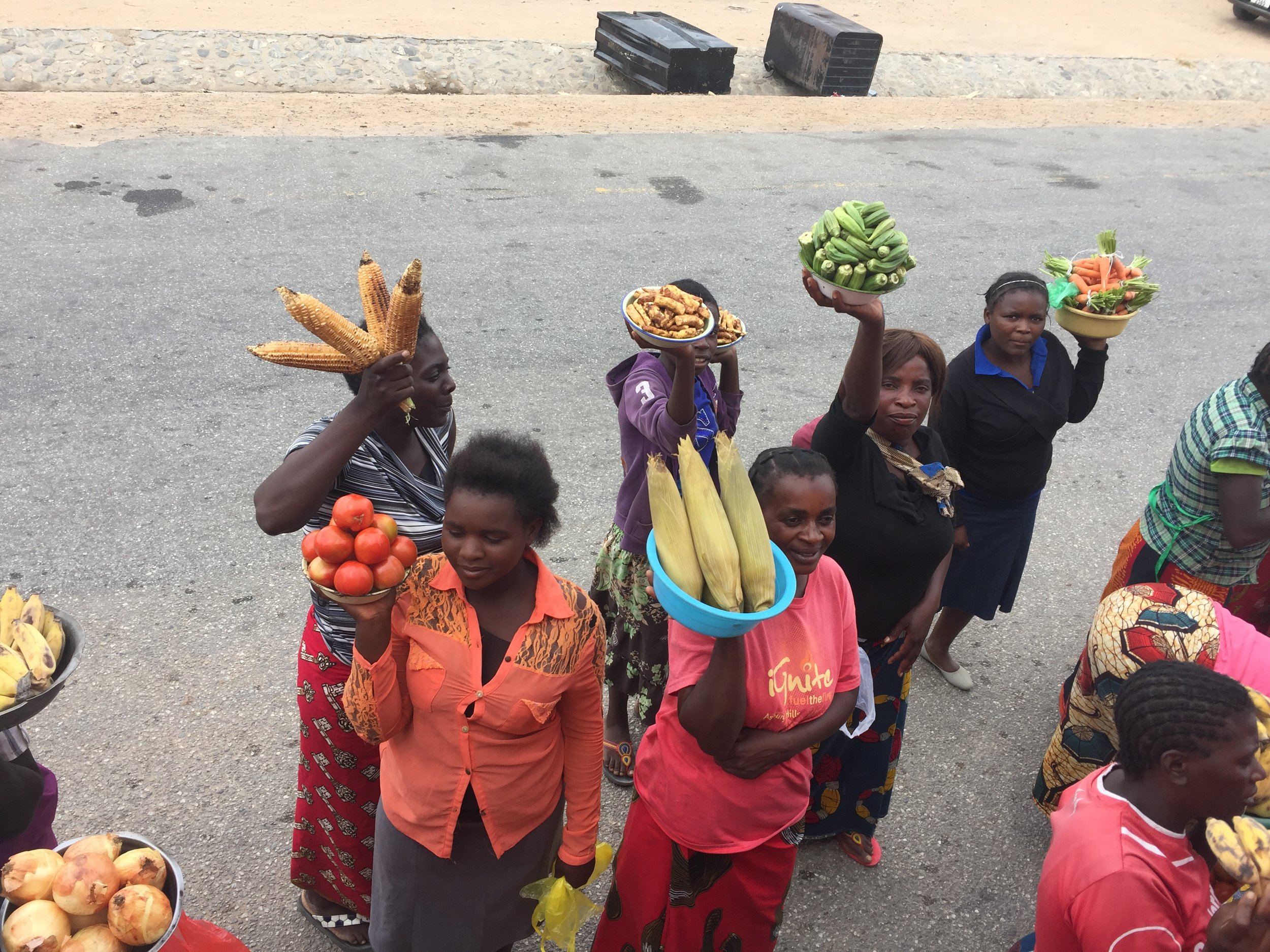 Women rush the side of the bus and reach up to sell fruits and vegetables to travelers.