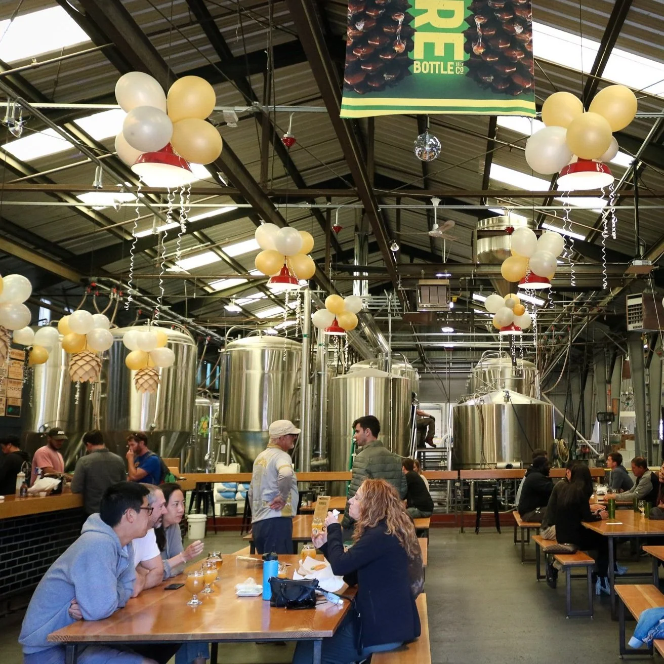 Barebottle's San Francisco Brewery & Taproom. Picture of people enjoying drinks in a large indoor warehouse. Seated at tables.