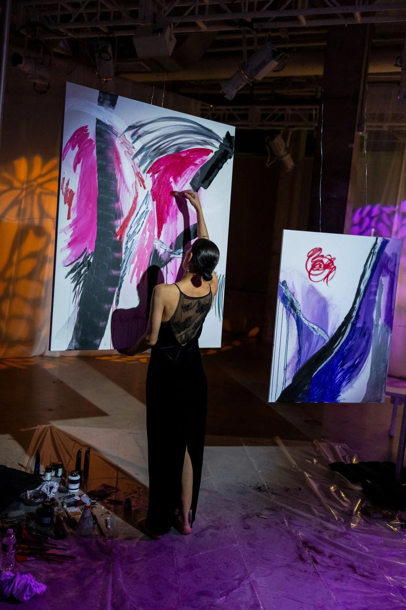 A woman in a black dress with a sheer back is painting abstract artwork on large canvases in a dimly lit studio or gallery. One canvas shows pink, black, and gray brushstrokes, while the other features purple, black, and red paint. Art supplies are on the floor nearby.
