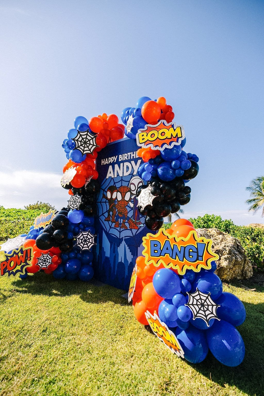 Superhero-themed birthday decoration with blue, black, red, and orange balloons, comic-style speech bubbles with words like "POW!", "BANG!", "BOOM", and "POW!" around a large vertical banner that reads "Happy Birthday Andy" featuring a cartoon spider character.