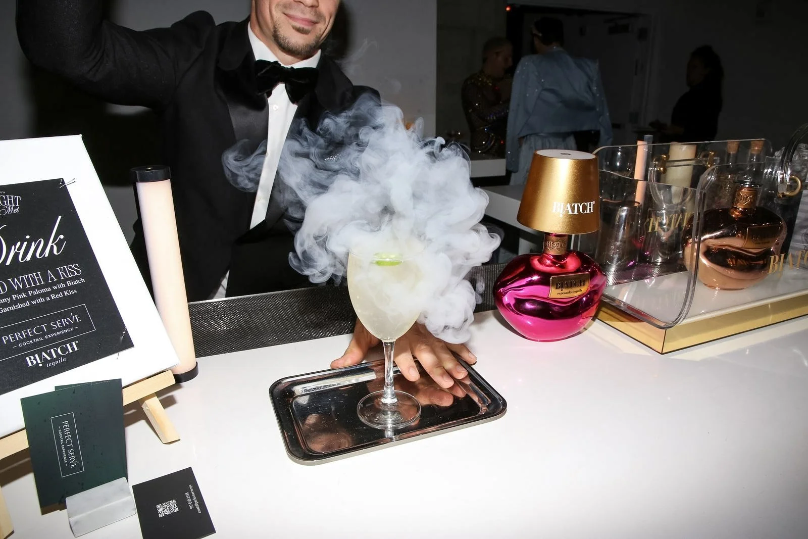 A bartender in a tuxedo preparing a dry ice cocktail at a bar, with smoke rising from the glass, surrounded by pink and rose gold perfume bottles and bar signs.
