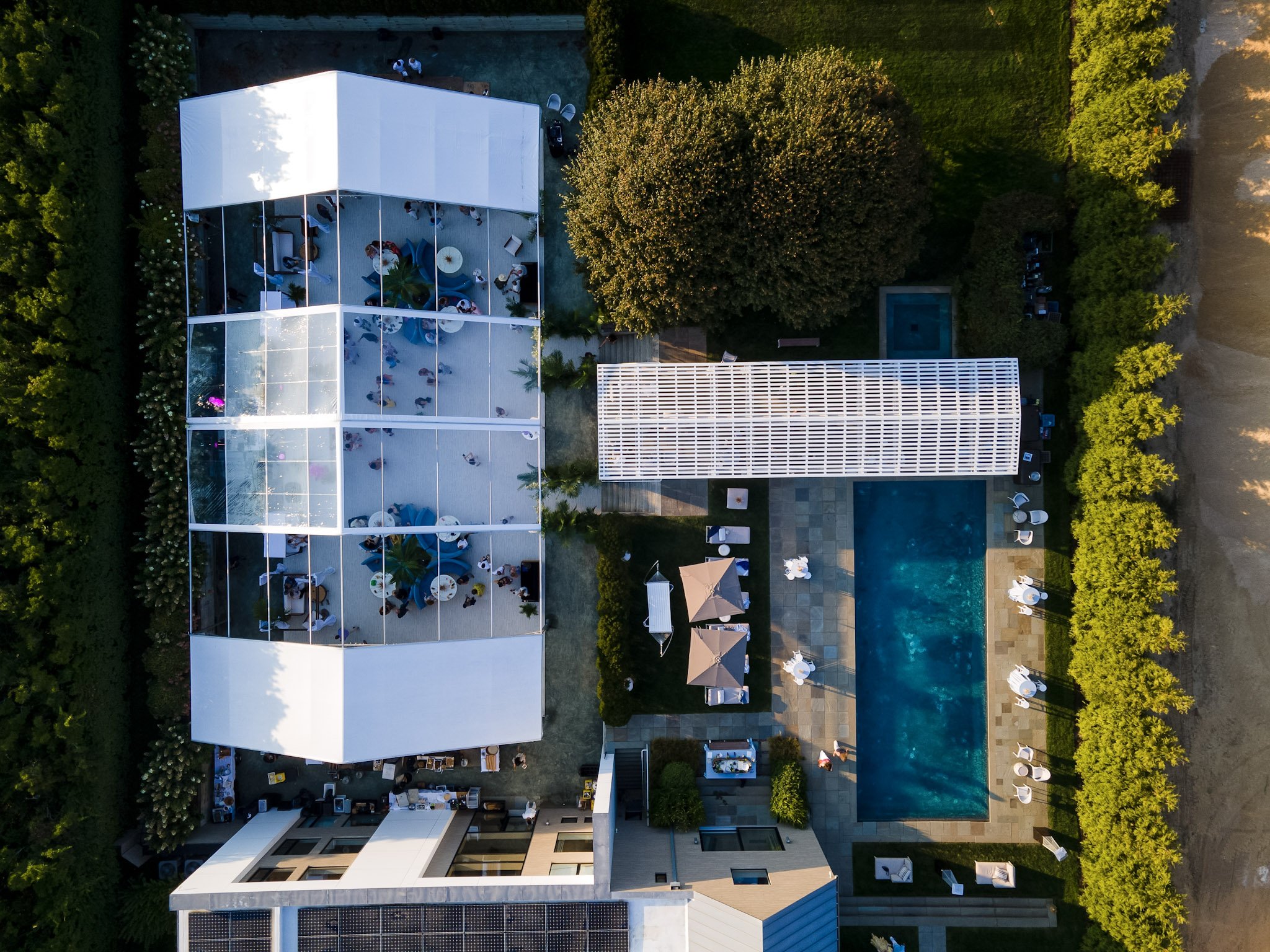 An aerial view of a modern outdoor pool area at sunset with lounging chairs, umbrellas, a pool, and a transparent enclosed space with people inside, surrounded by greenery.